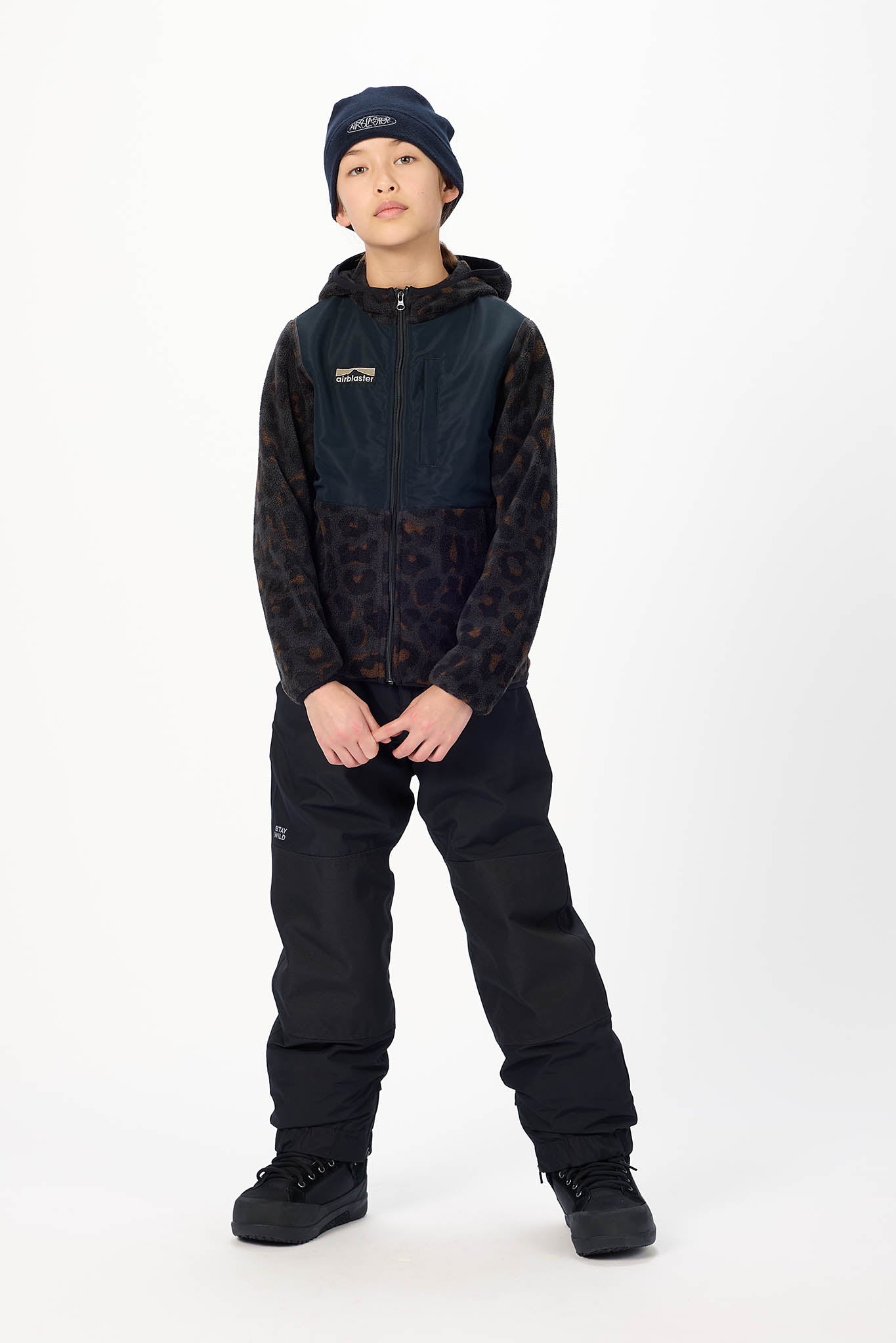 A child model wearing a dark navy and black patterned hooded jacket, black snow pants, black boots, and a navy beanie, standing against a plain white studio background.