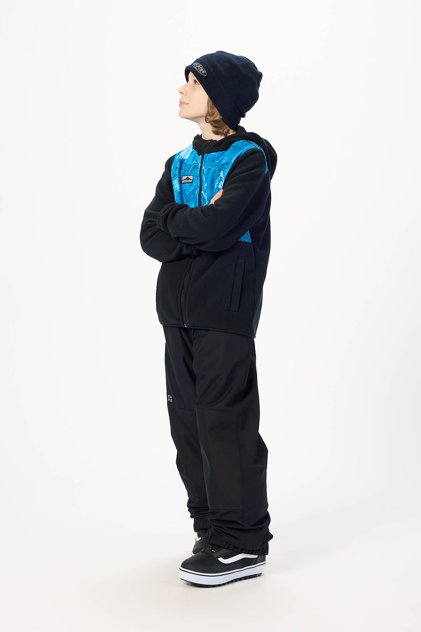 A child model wearing a black and blue hooded jacket, black pants, a black beanie, and black sneakers, standing in profile against a plain white studio background.