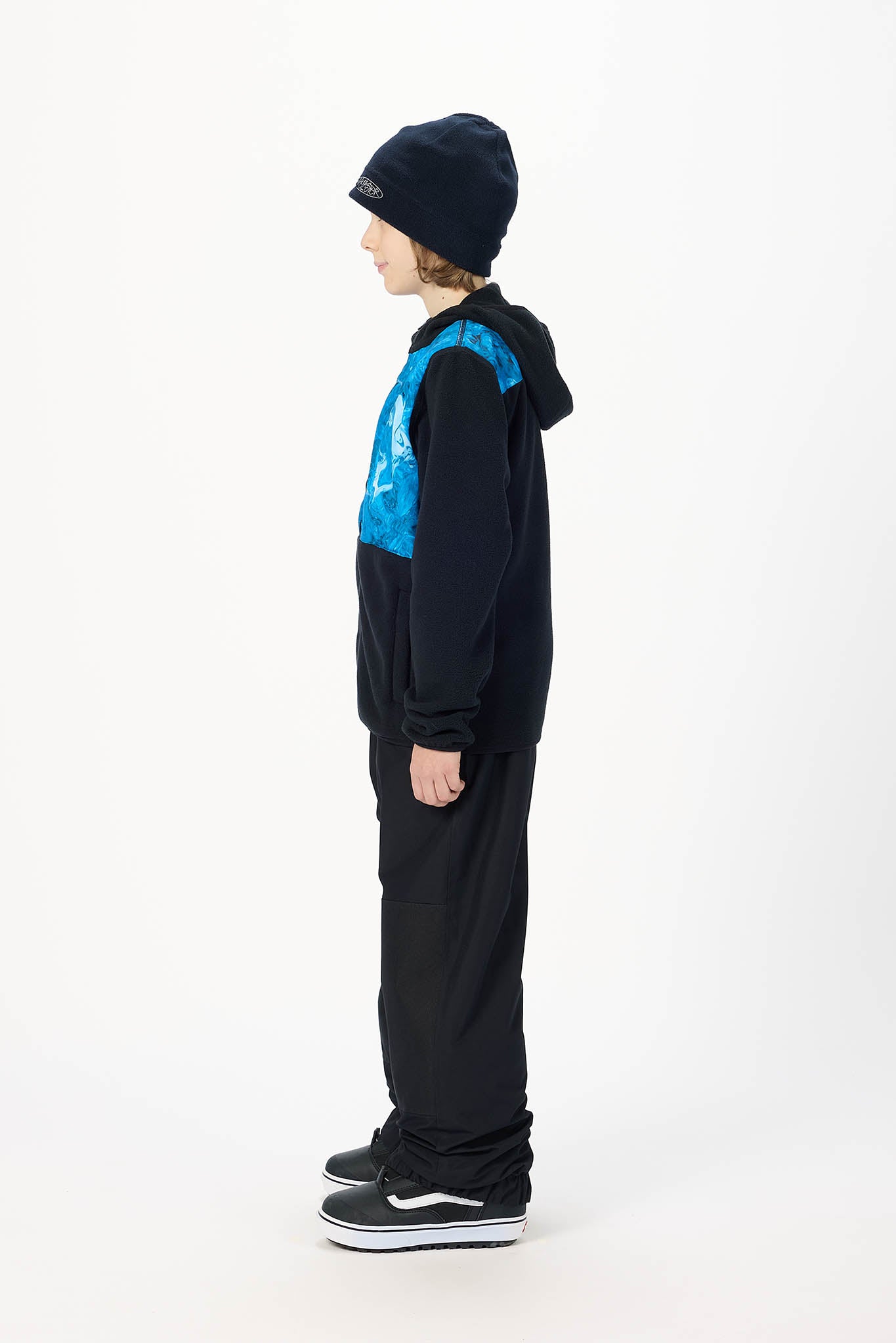 A child model shown in profile wearing a black hooded sweatshirt with a bright blue graphic chest panel, black pants, black sneakers, and a black beanie on a plain white studio background.