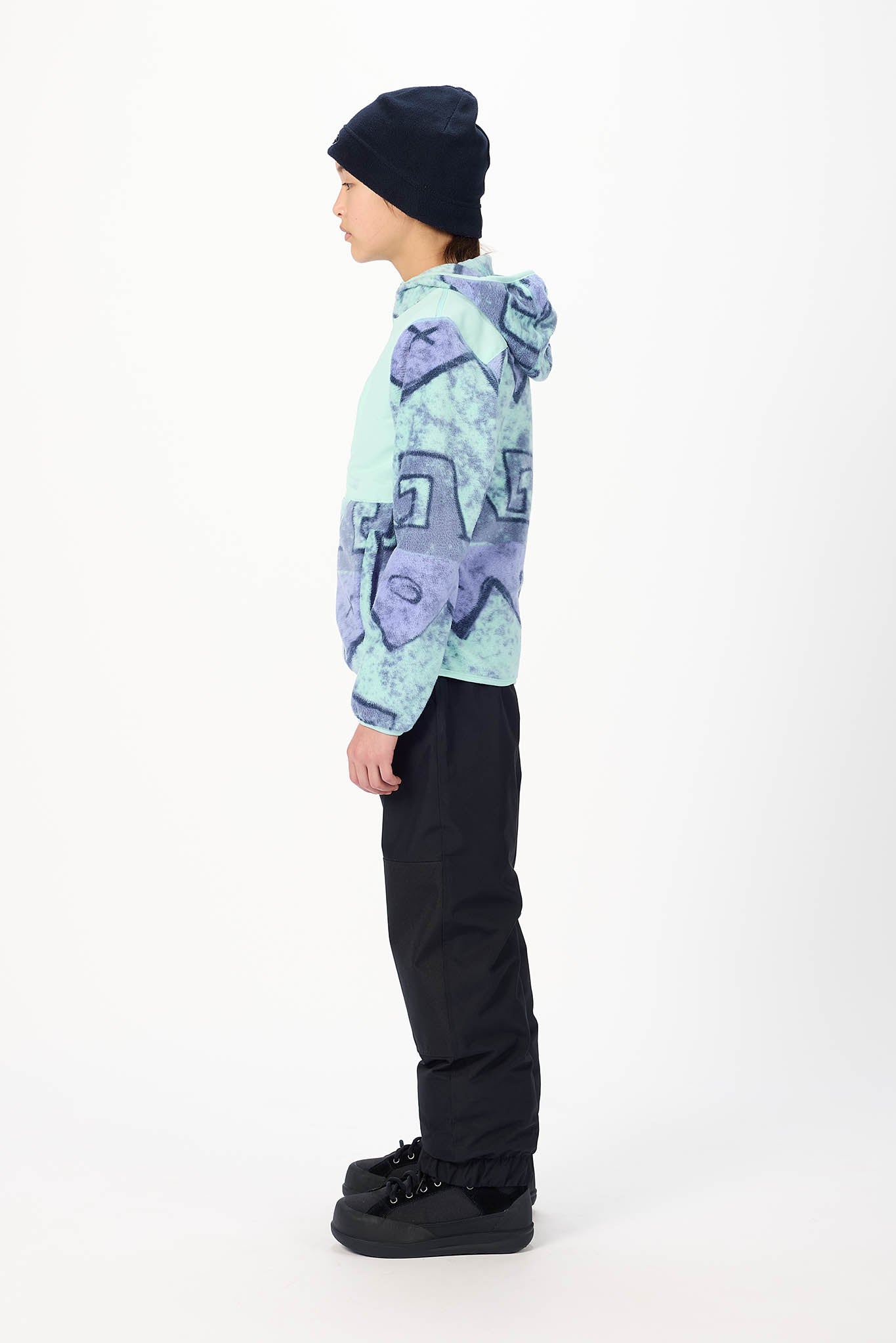 A child standing in profile wearing a light blue and purple patterned hoodie, black pants, black shoes, and a black beanie against a plain white studio background.