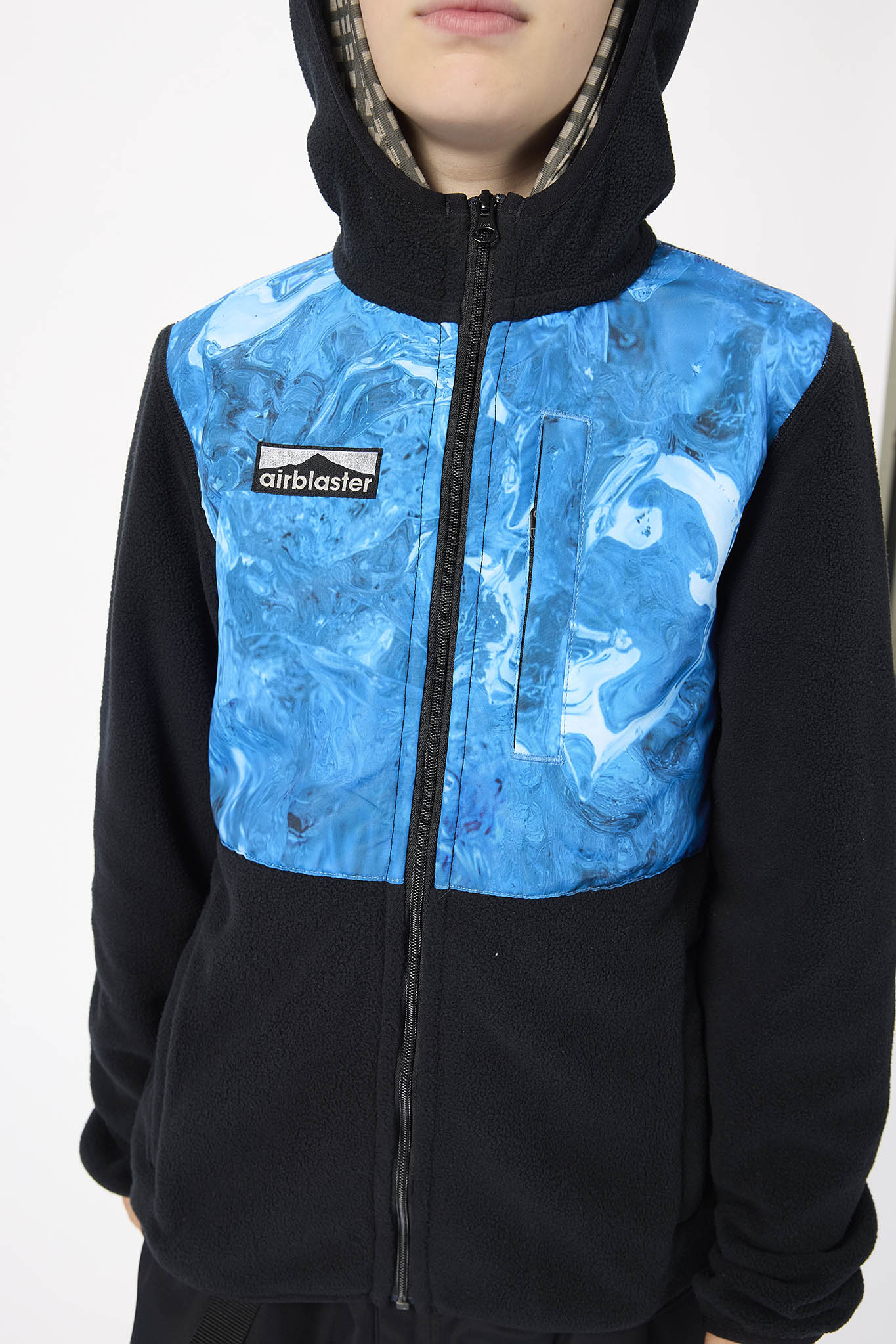 A close-up of a person wearing a zip-up hooded jacket with a black body and sleeves, and a bright blue marbled pattern across the chest. A small 'airblaster' logo patch is visible on the left chest.