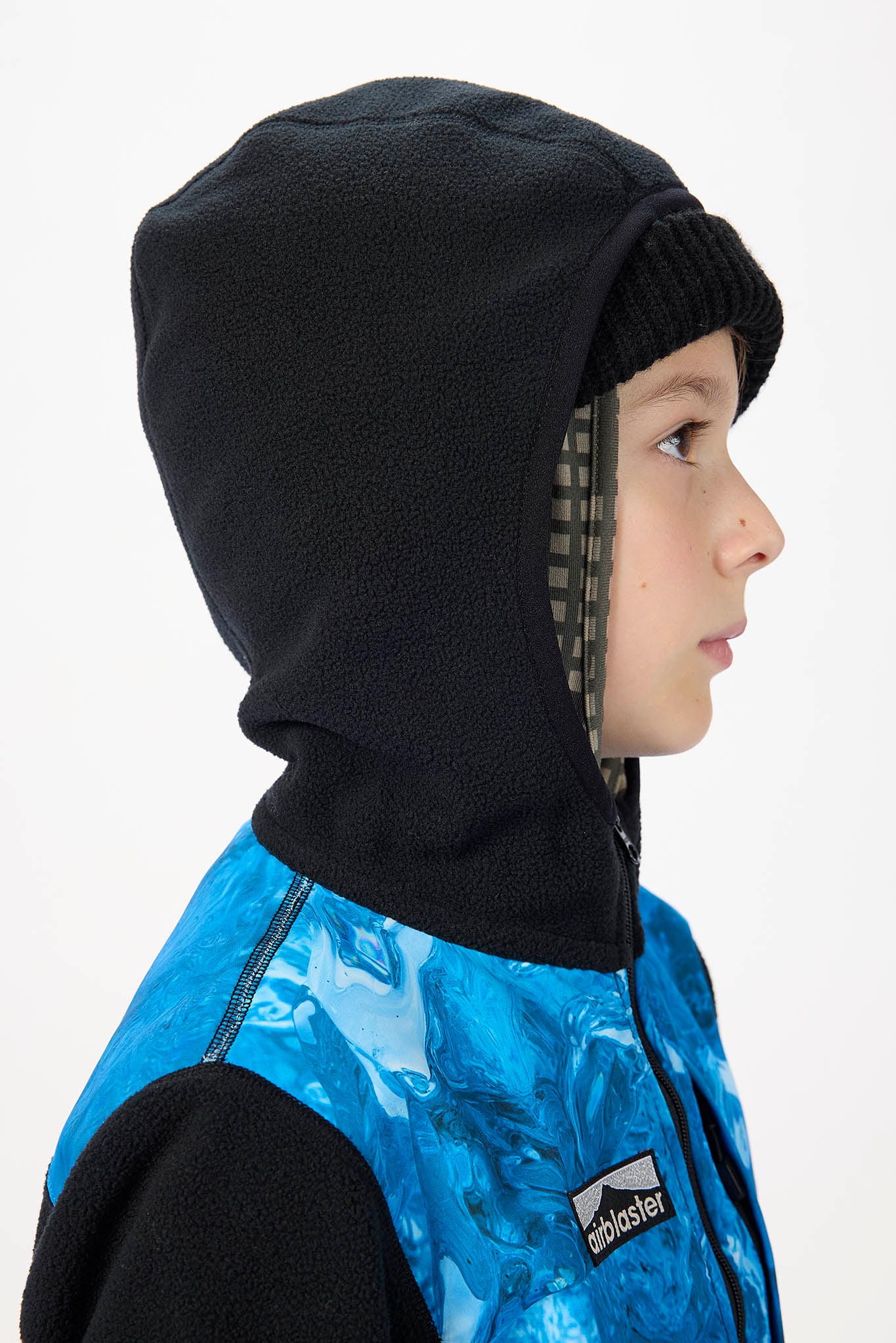 A child wearing a black fleece hooded balaclava over a blue and black patterned zip-up jacket, shown in profile against a white background.