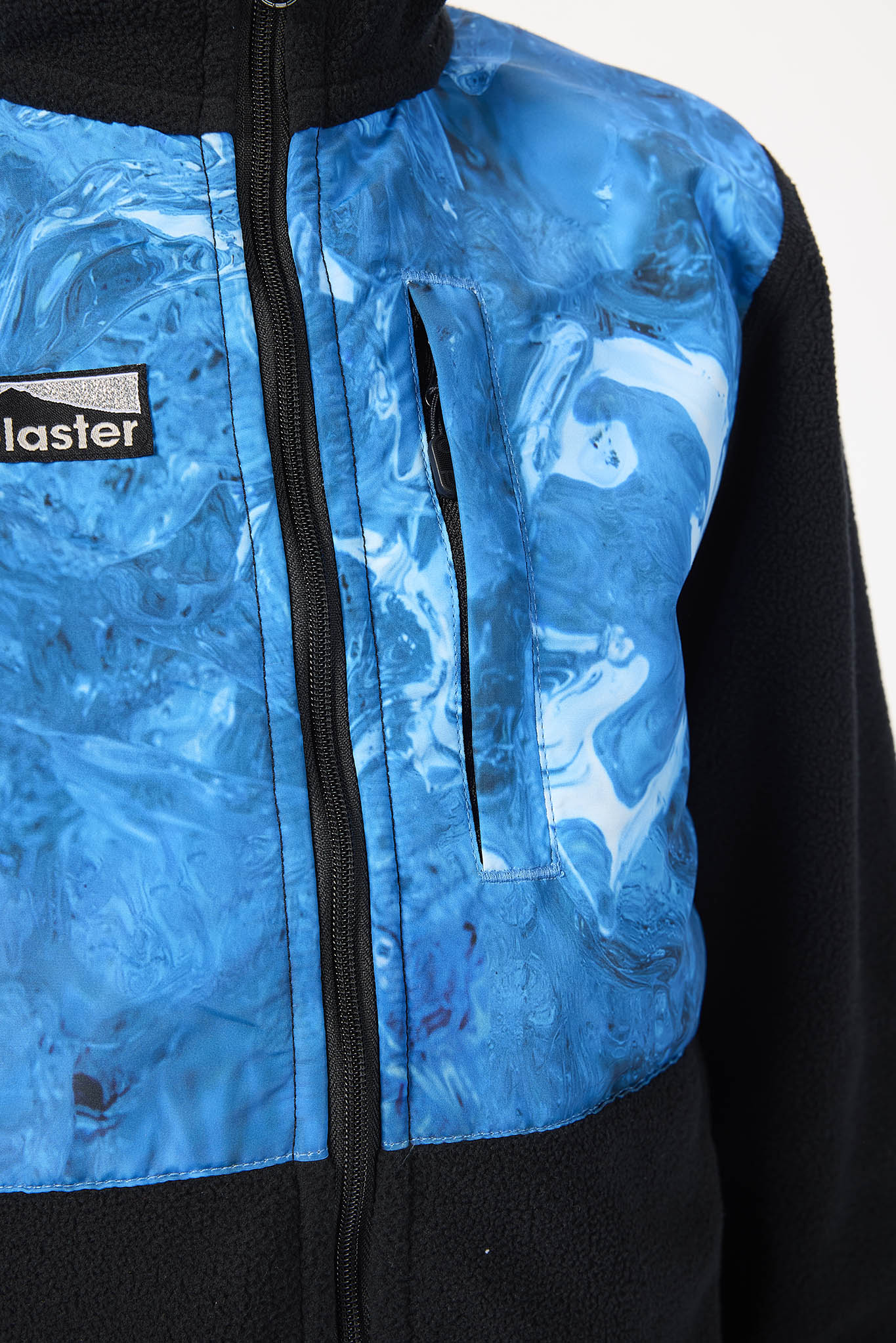 Close-up of a zip-front jacket with a blue marbled pattern on the front panel and black sleeves, plus a visible 'blaster' logo patch on the chest.