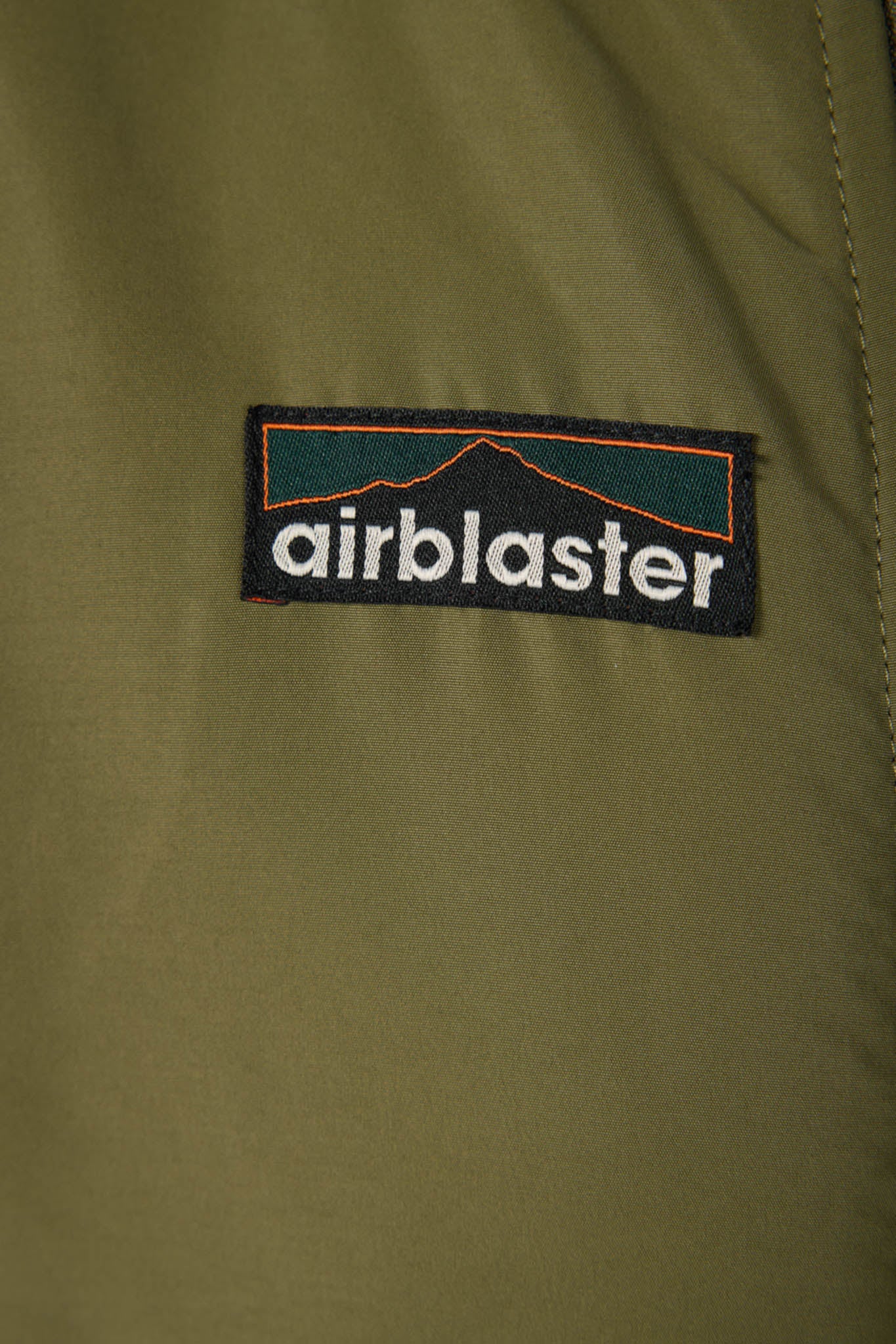 Close-up of an olive green jacket or fabric panel featuring a rectangular black Airblaster logo patch with white text and a dark teal and orange graphic.