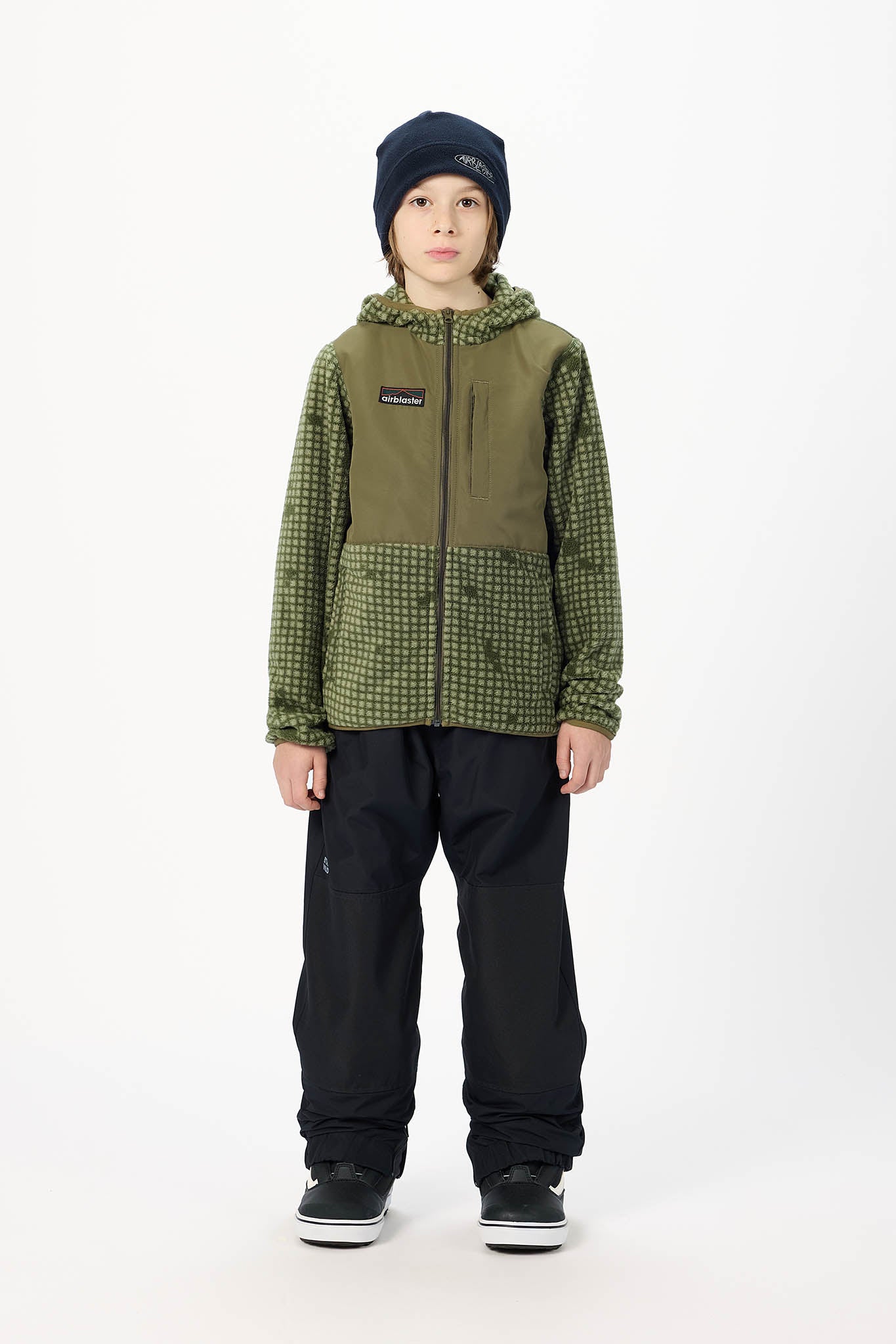 A child model wearing a green and black patterned hooded jacket, black snow pants, a dark navy beanie, and black sneakers, standing against a plain white studio background.