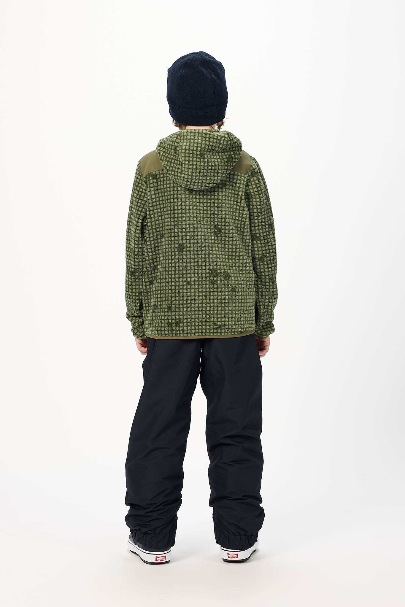 A child shown from the back wearing an olive green and black patterned hooded jacket, black snow pants, a black beanie, and white sneakers, standing against a plain white studio background.