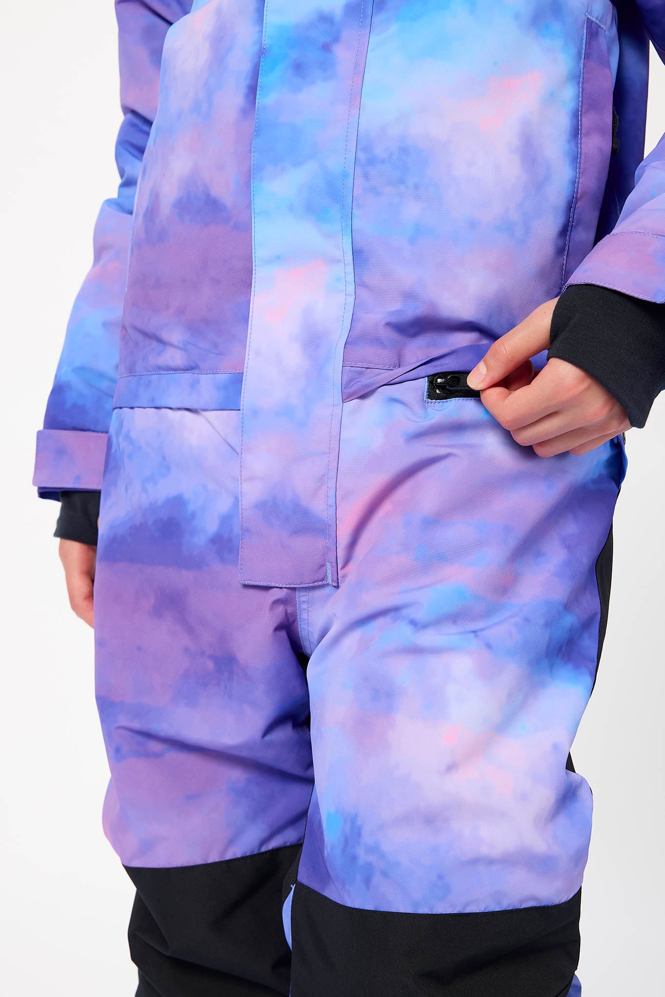 Close-up of a person wearing a purple and blue tie-dye snowsuit with black cuffs and black lower legs, shown against a plain light background.