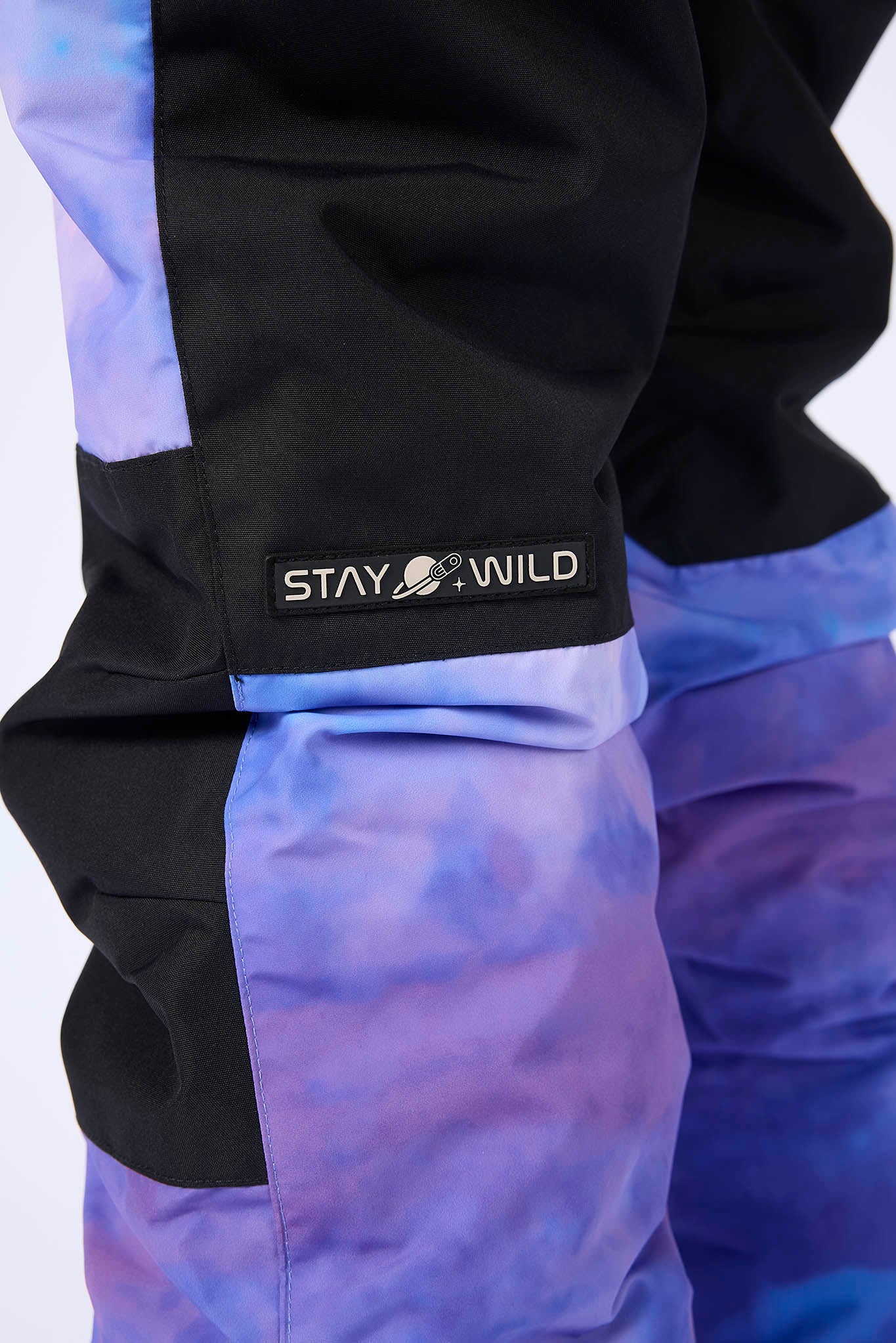 Close-up of a pair of color-blocked ski pants featuring black upper sections and purple-to-blue tie-dye lower sections, with a black cuff band displaying the 'STAY WILD' logo.