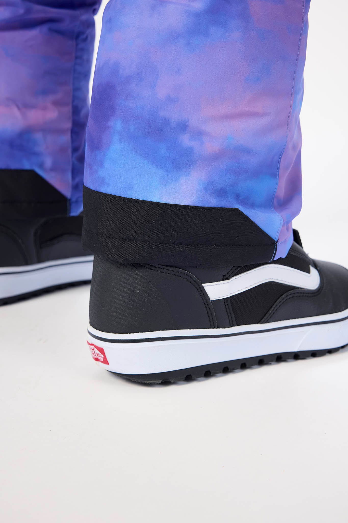 Close-up of a person wearing black Vans high-top sneakers with white side stripes and white soles, paired with blue and purple tie-dye pants, against a light gray studio background.