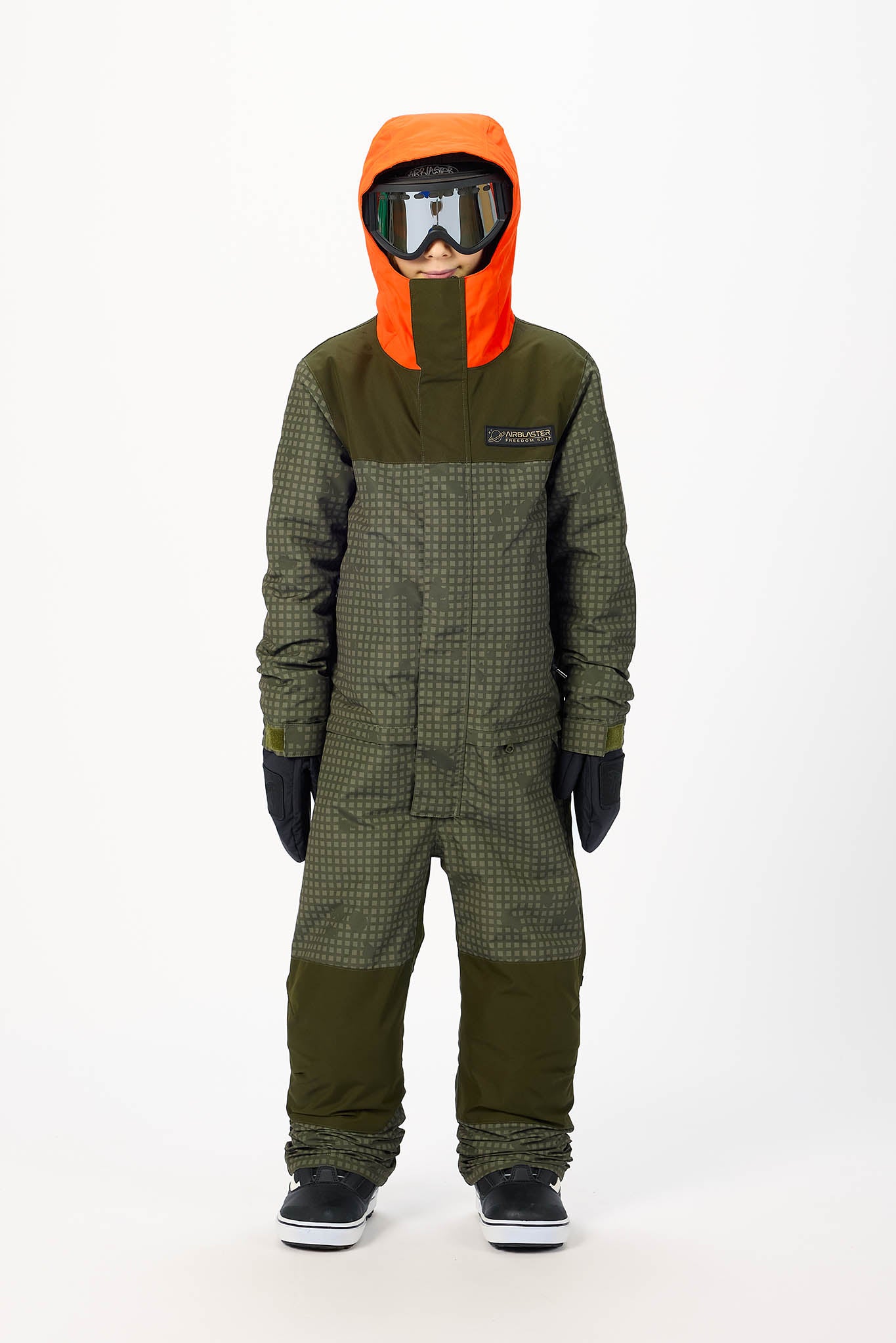 A child modeling a full-body snowsuit with an attached hood, bright orange hood, and olive green body with a subtle grid pattern. The child is also wearing ski goggles, black gloves, and black-and-white sneakers against a plain white studio background.