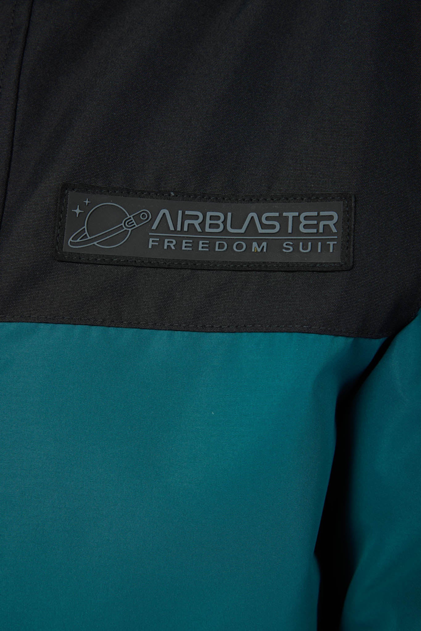Close-up of a color-blocked jacket or suit with a black upper section and teal lower section, featuring a rectangular patch that reads 'AIRBLASTER FREEDOM SUIT' with a small planet graphic.