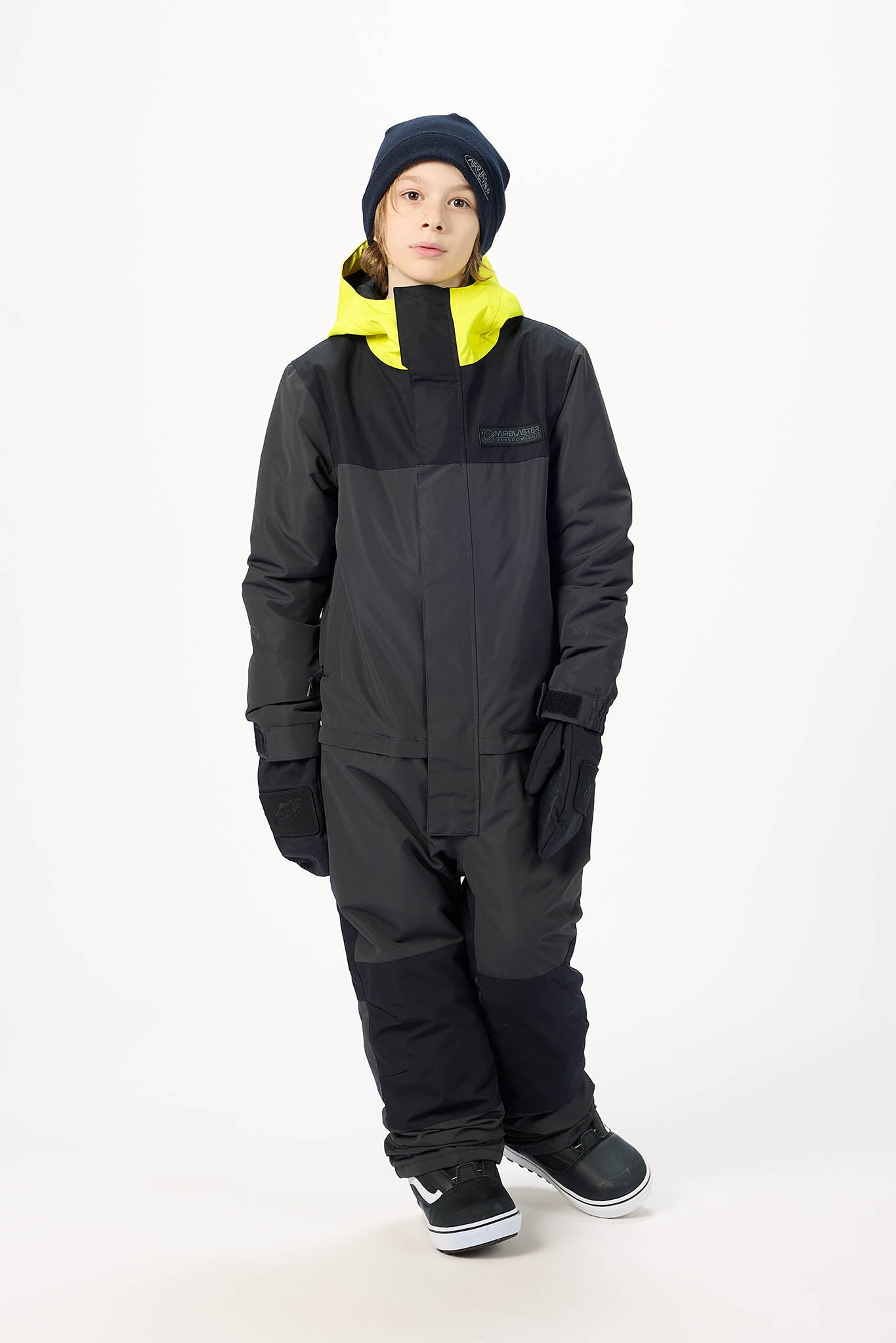 A child wearing a black snowsuit with a bright yellow hood lining, a black beanie, and black sneakers, standing against a plain white studio background.