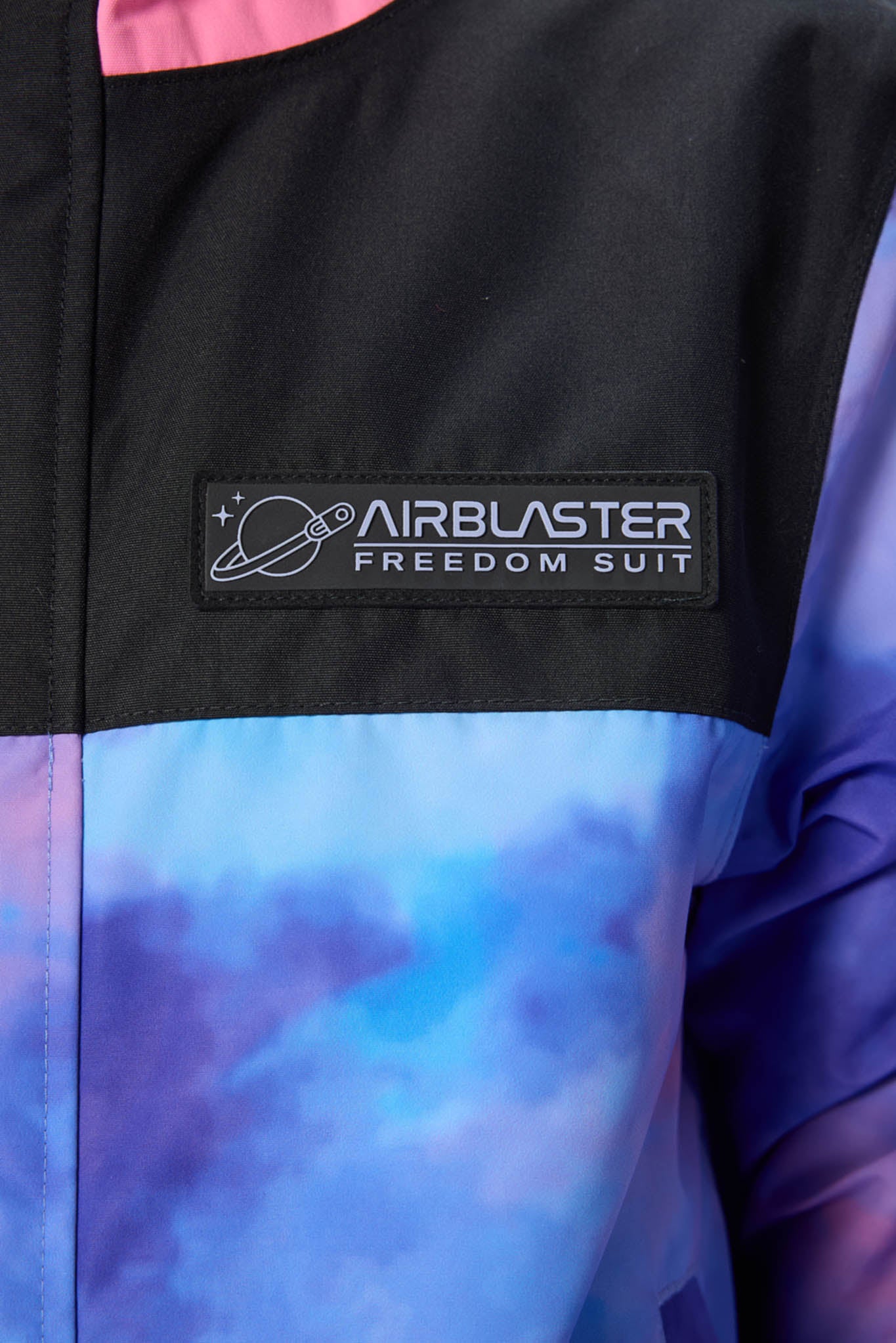 Close-up of a colorblock jacket featuring a black upper chest with a 'AIRBLASTER FREEDOM SUIT' patch, and a blue and purple tie-dye patterned lower section.