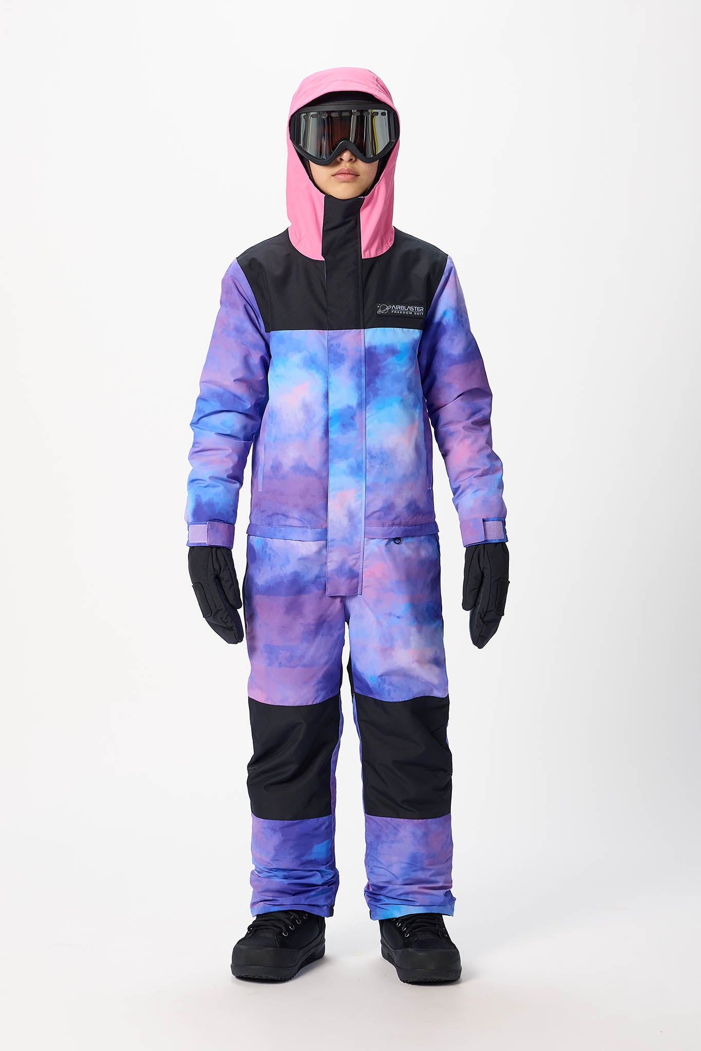 A child wearing a hooded snowsuit with a purple and blue galaxy print, black chest and lower-leg panels, black gloves, black boots, and dark goggles, standing against a light gray studio background.