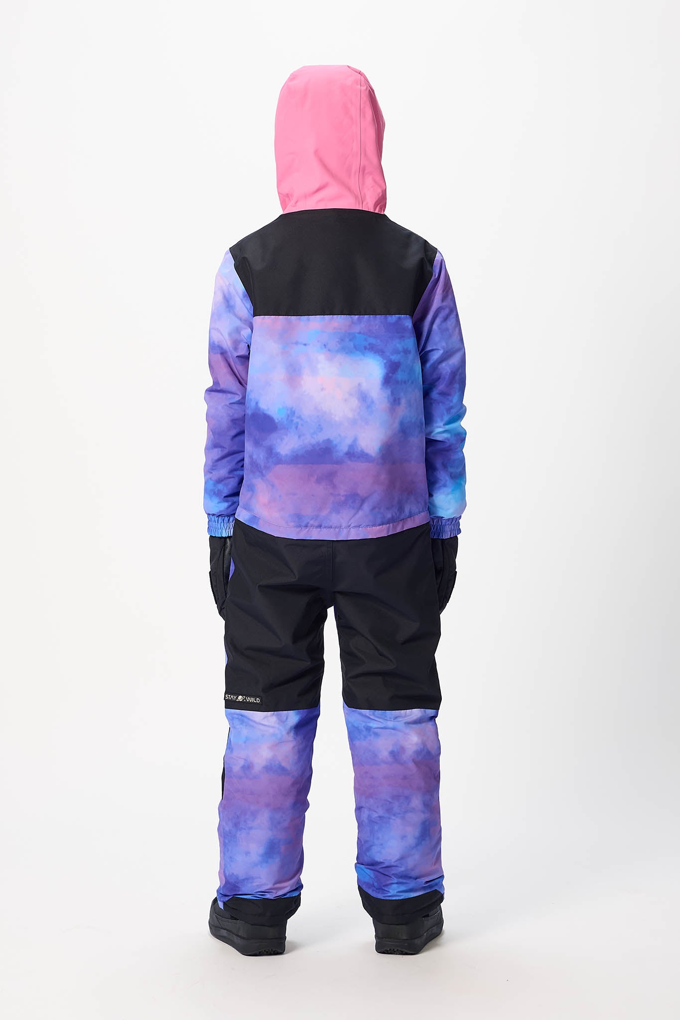 A child viewed from the back wearing a hooded snowsuit with a pink hood, black upper back and shoulders, and a blue-purple cloud-like print on the sleeves and back, paired with black pants and black boots on a light gray studio background.