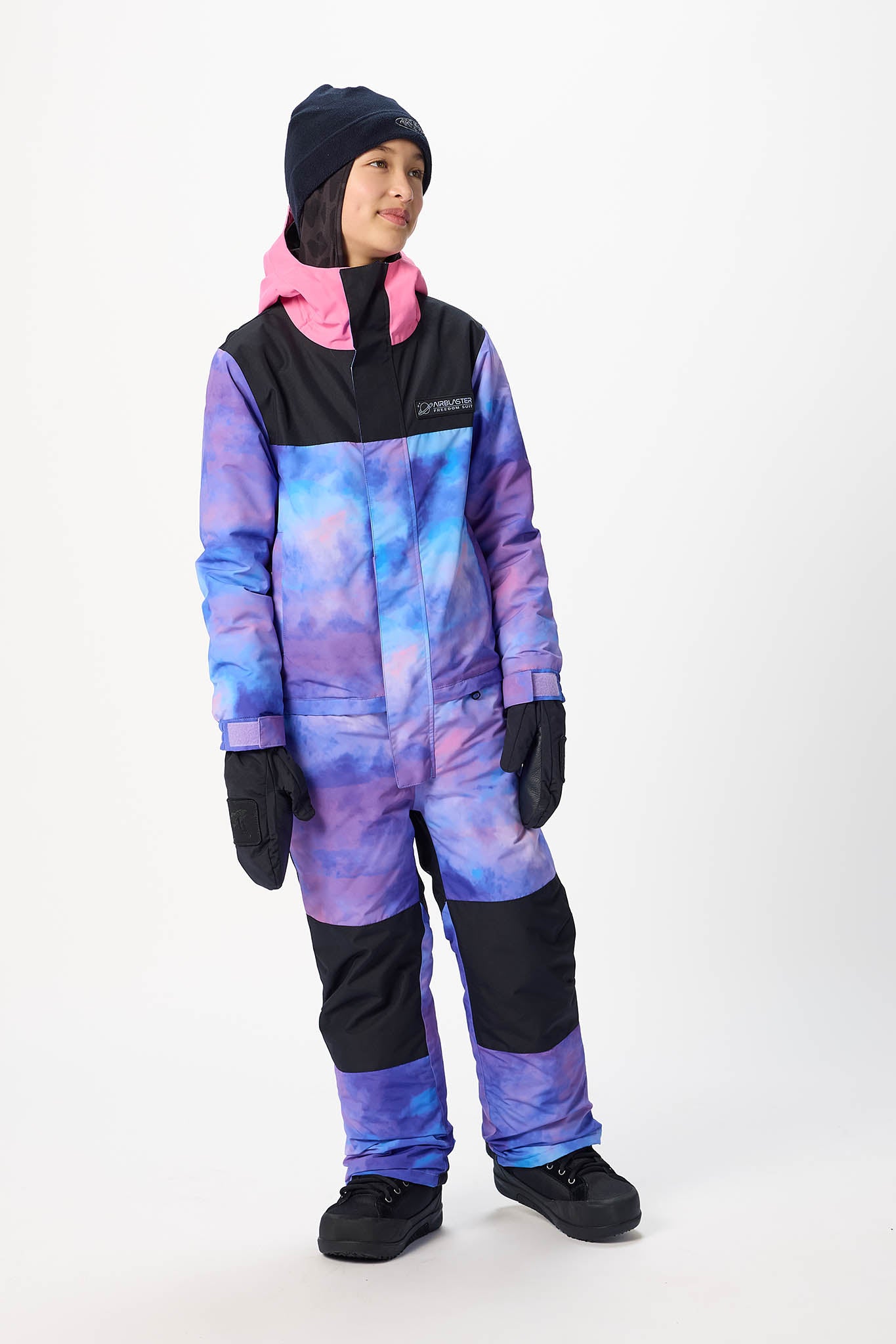 A child wearing a full-body snowsuit with a purple and blue galaxy print, black panels, and a pink hood, standing against a plain white studio background.