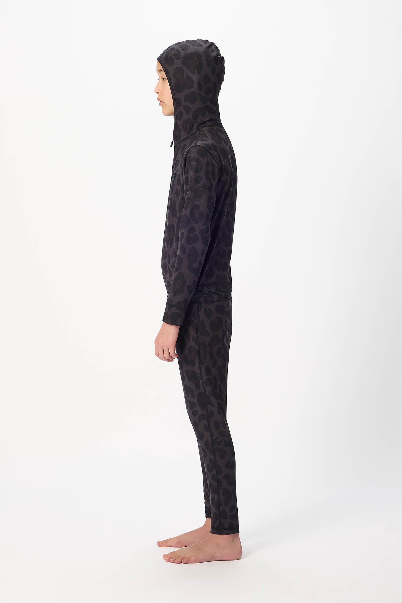 A child model shown in profile wearing a full-body black hooded one-piece outfit with an allover tonal leopard-like pattern, standing barefoot against a plain light gray studio background.