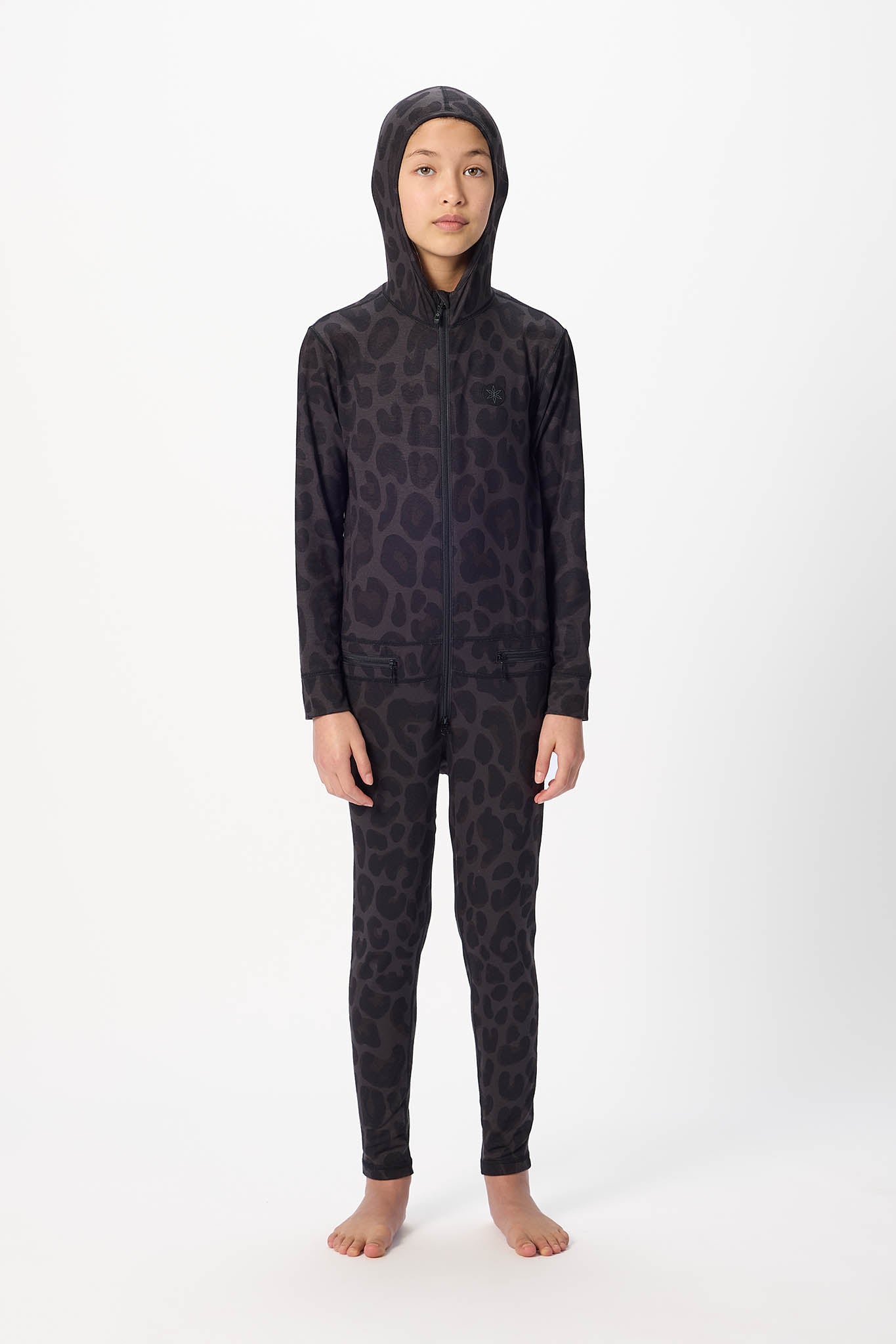 A child model wearing a full-body hooded one-piece swimsuit or wetsuit with a dark leopard print pattern, standing against a plain white studio background.