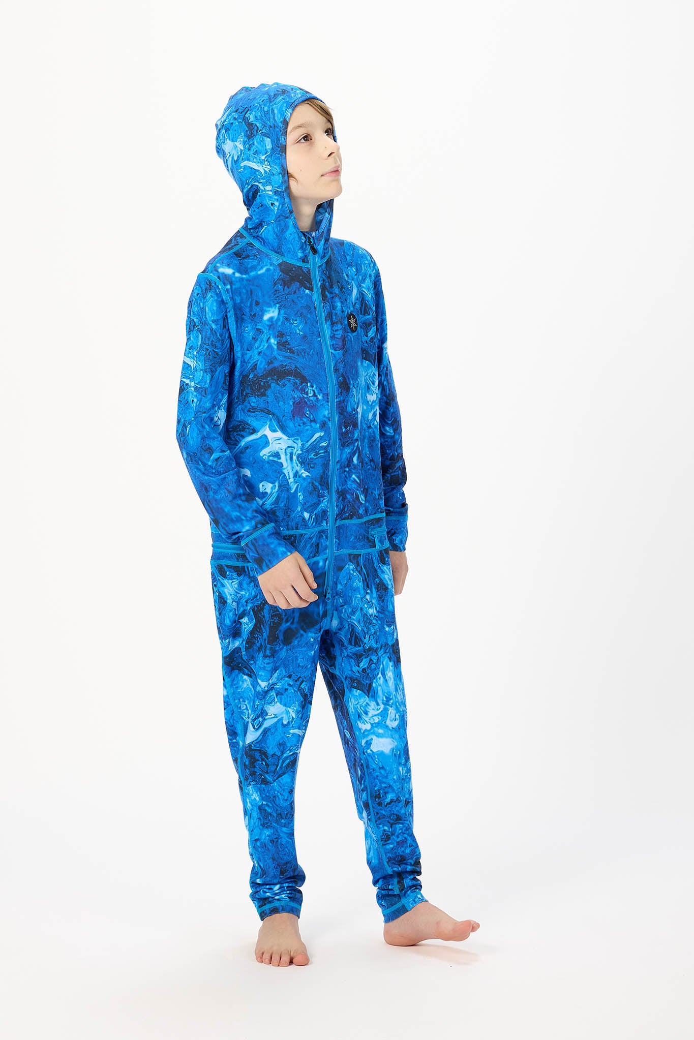 A child wearing a full-body hooded one-piece jumpsuit in a bright blue and navy abstract pattern, standing against a plain white studio background.