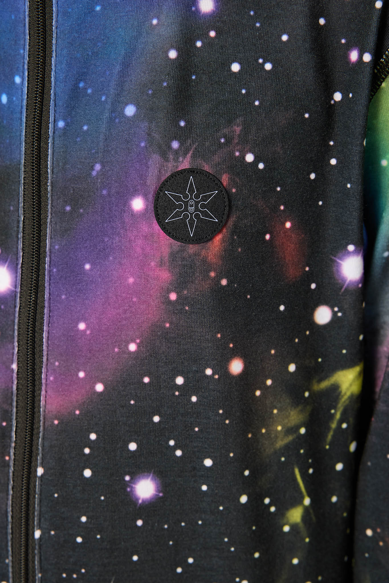 Close-up of a black zippered jacket featuring a vibrant galaxy print with purple, pink, and blue nebulae and white stars. A circular black patch with a white geometric star design is stitched on the chest.