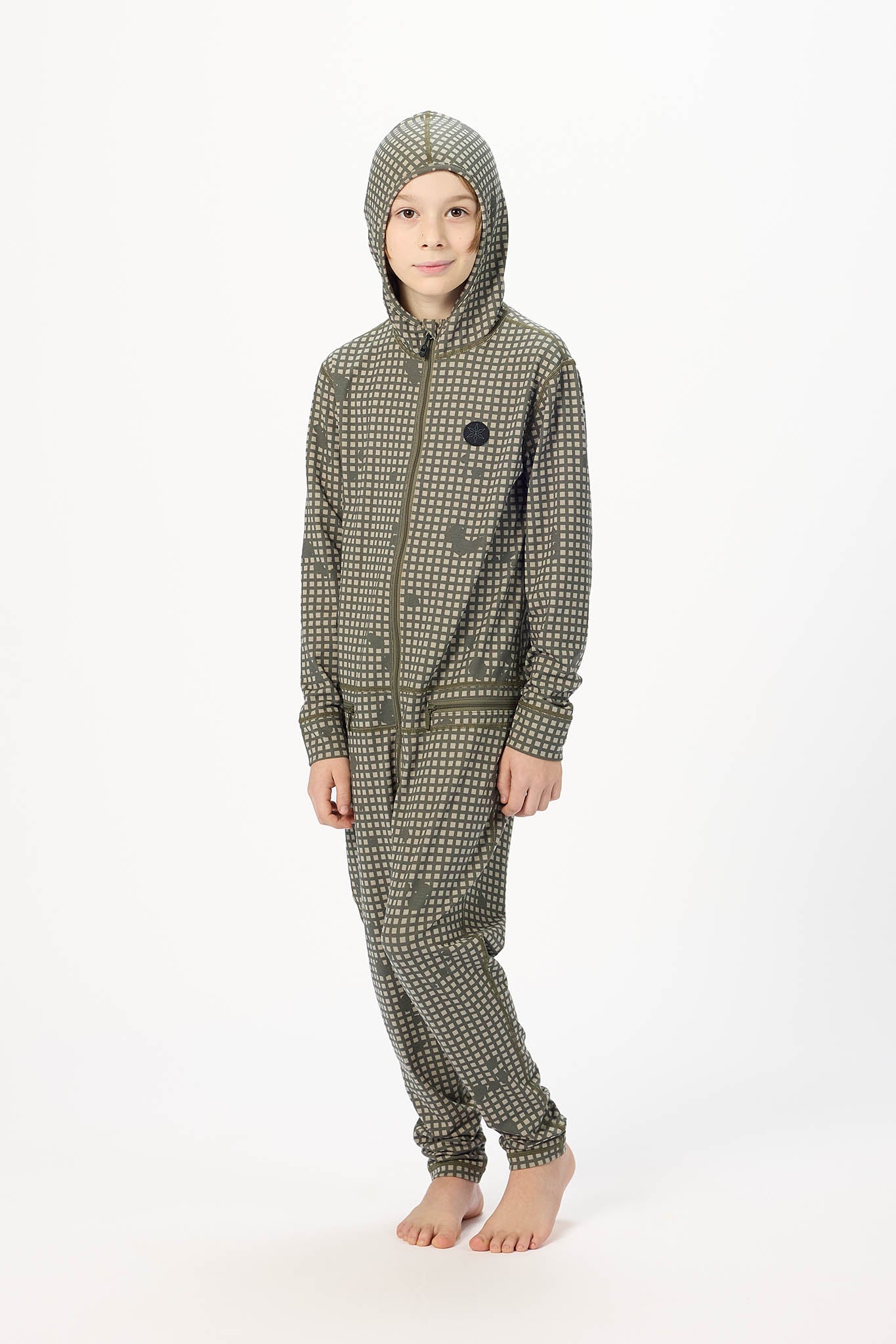 A child wearing a hooded one-piece jumpsuit with a small-scale green and white pattern, standing against a plain white studio background.