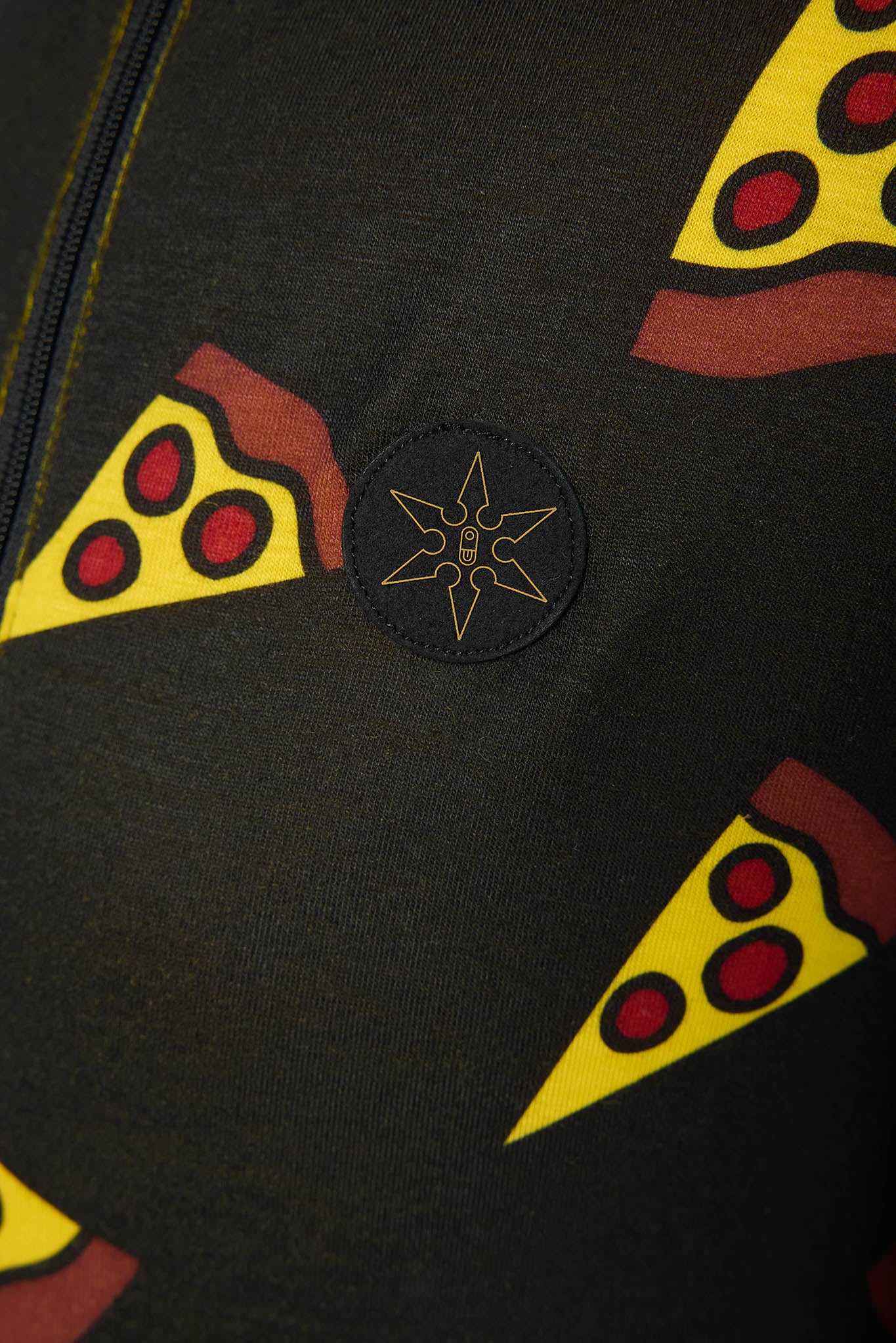 Close-up of a black fabric garment featuring a repeating yellow pizza slice pattern with red toppings and a central circular black patch with a gold star-like emblem.