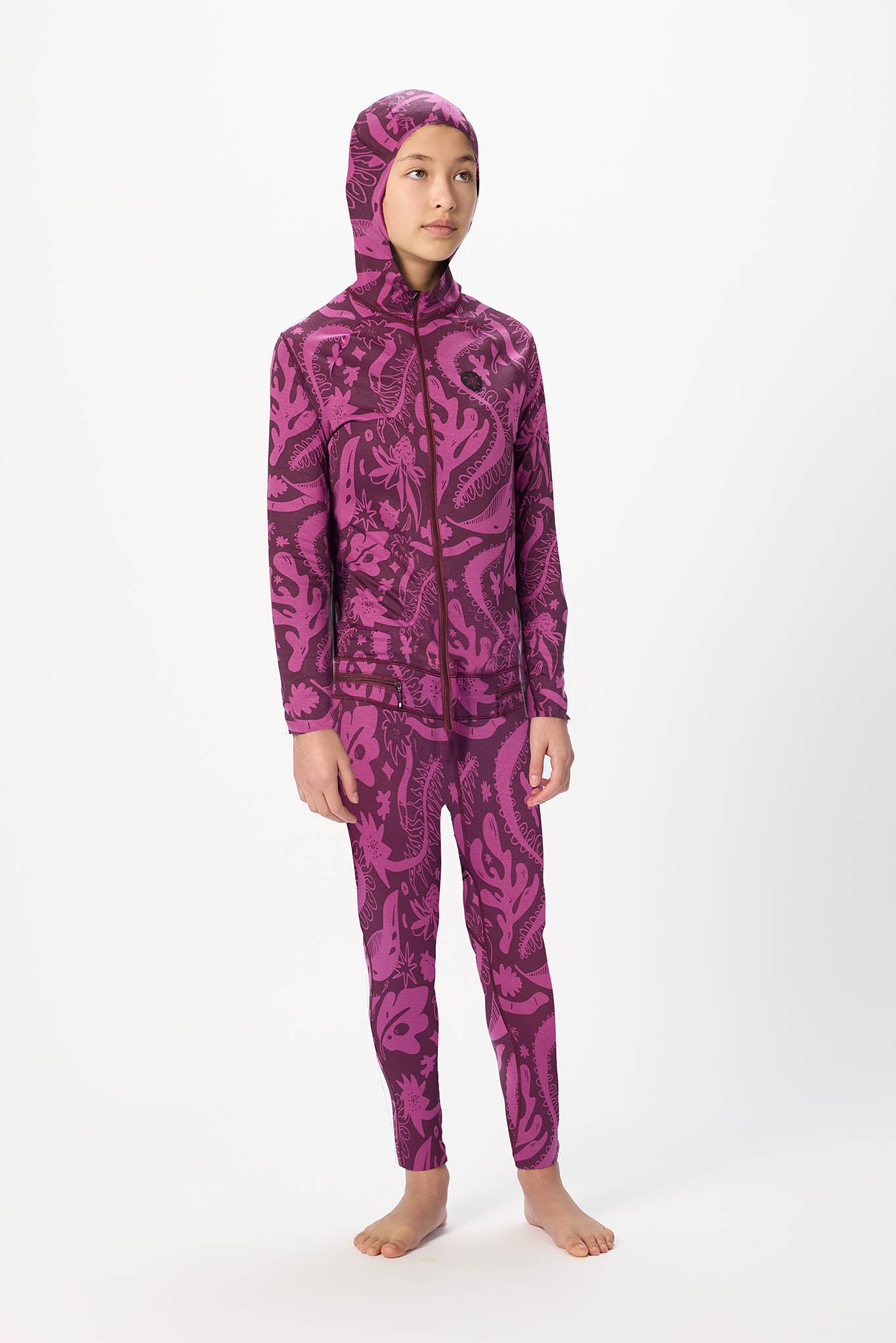 A child model wearing a matching two-piece tracksuit with a hooded jacket and pants, featuring a bold magenta and dark purple abstract pattern, set against a plain white studio background.