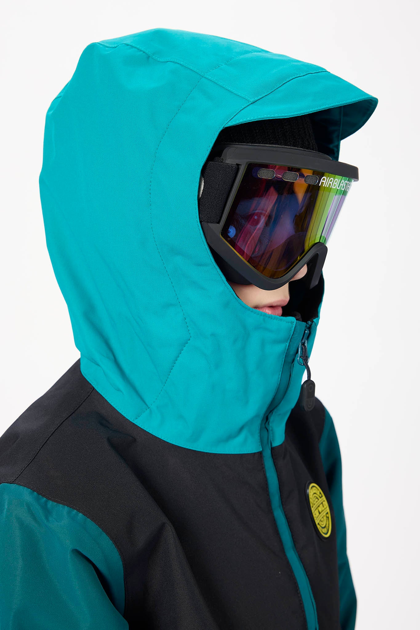 A close-up side profile of a person wearing a teal and black ski jacket with a matching hood, black ski goggles, and a small yellow circular logo patch on the chest.