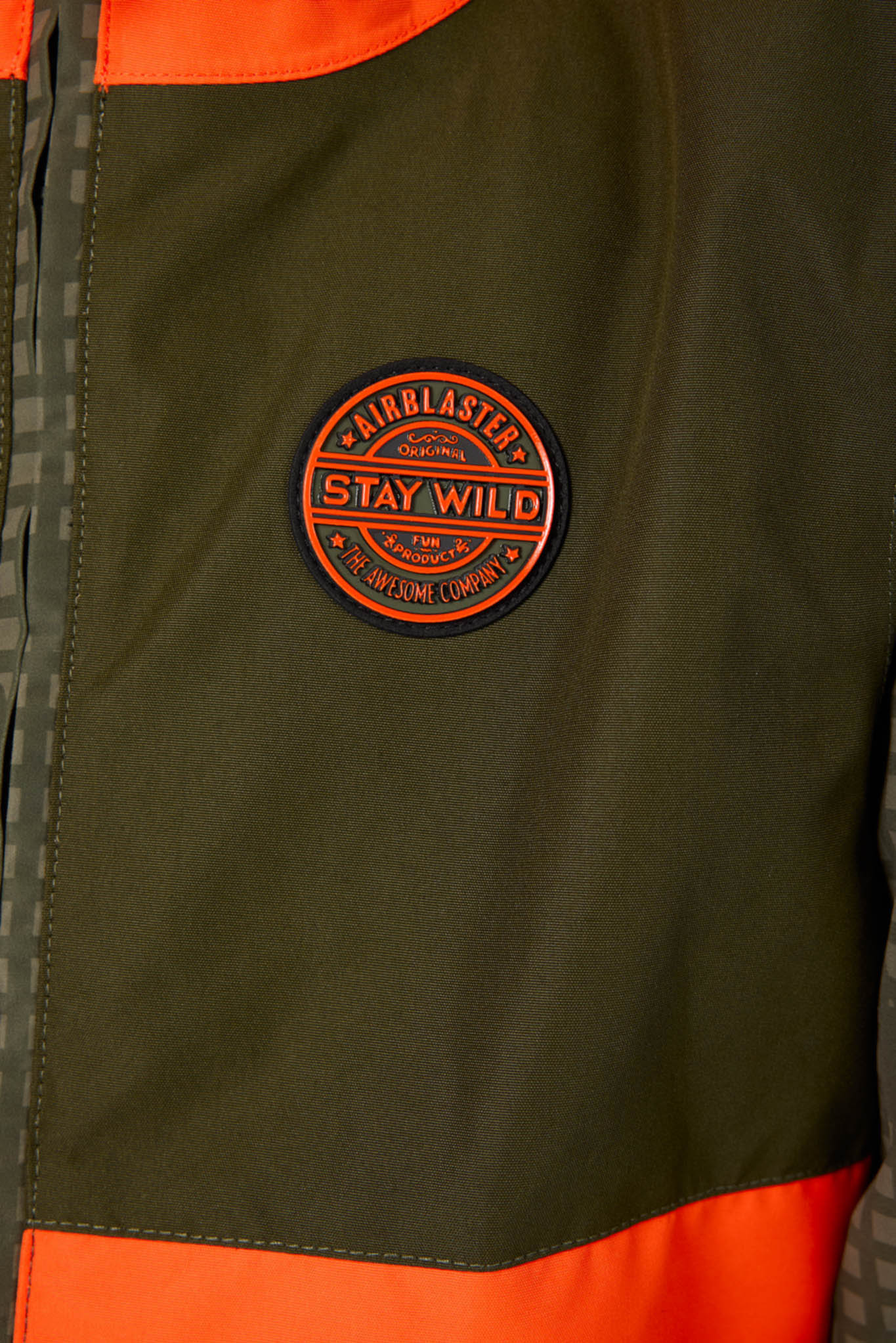 Close-up of a dark olive green jacket with bright orange contrast panels and a circular orange and black 'Airblasters Stay Wild' patch on the chest.