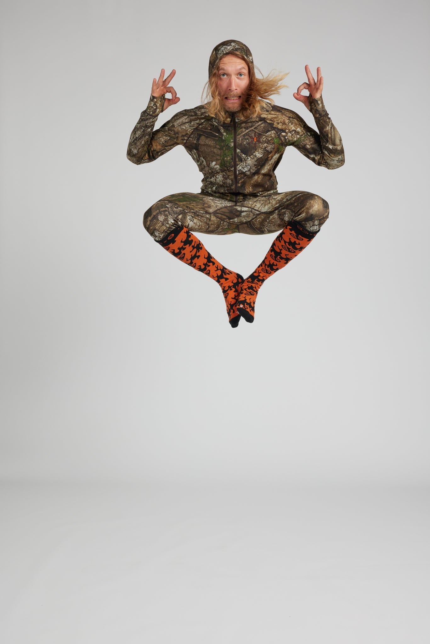 A person mid-jump against a light gray studio background, wearing a full-body camouflage outfit with bright orange and black patterned socks.