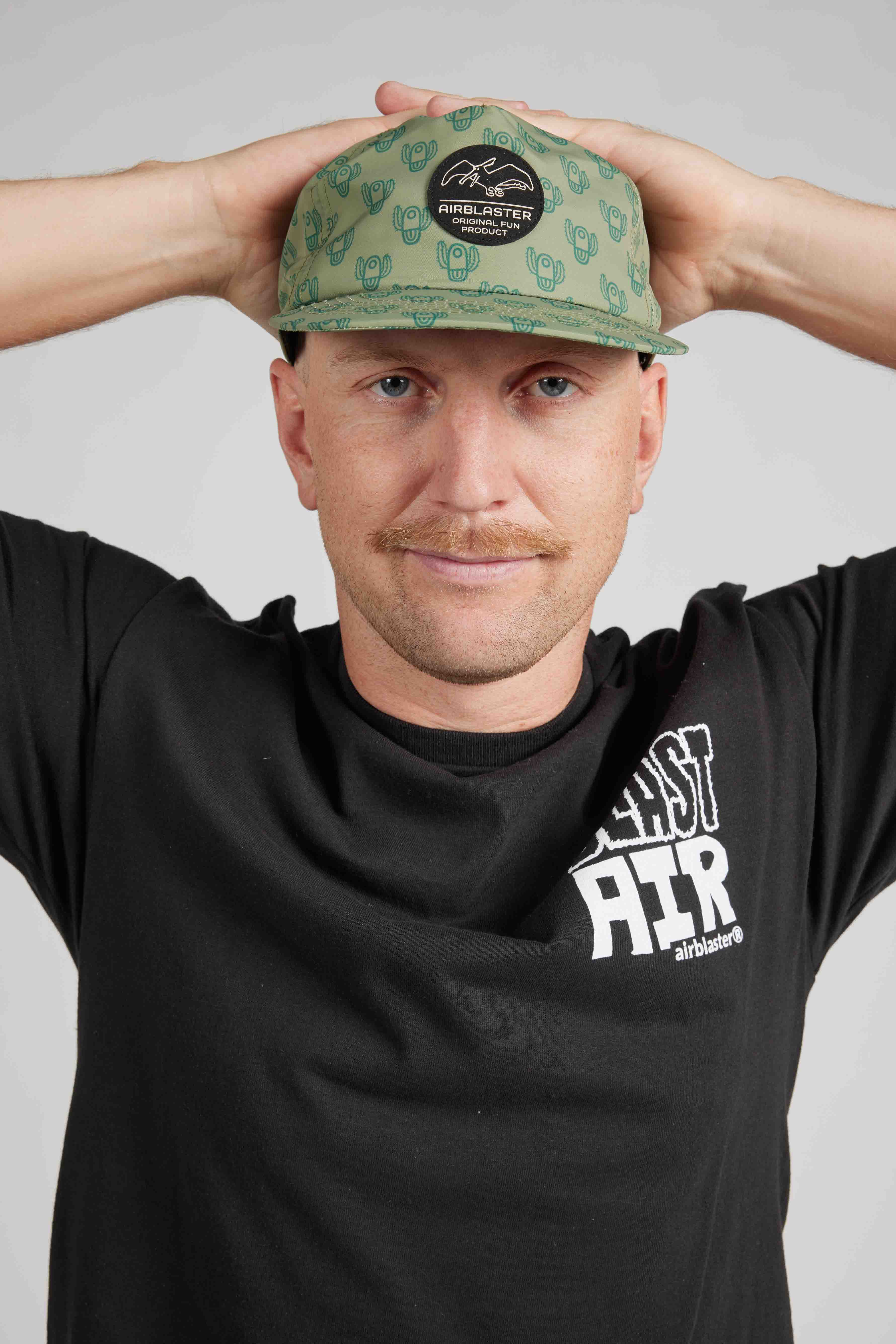 Man wearing a black t-shirt with a logo and a green cap with a pattern, against a plain background