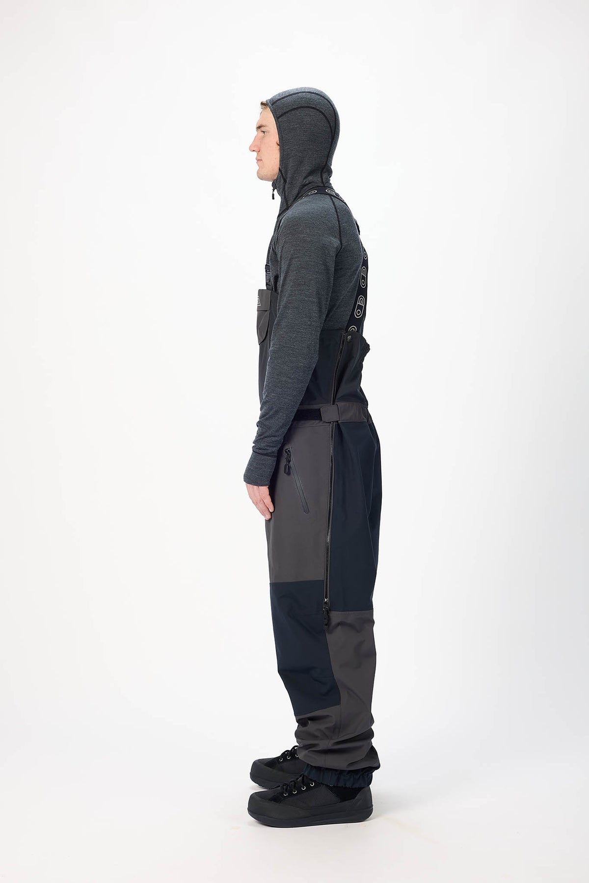 Person wearing a dark gray hooded outfit with suspenders on a white background
