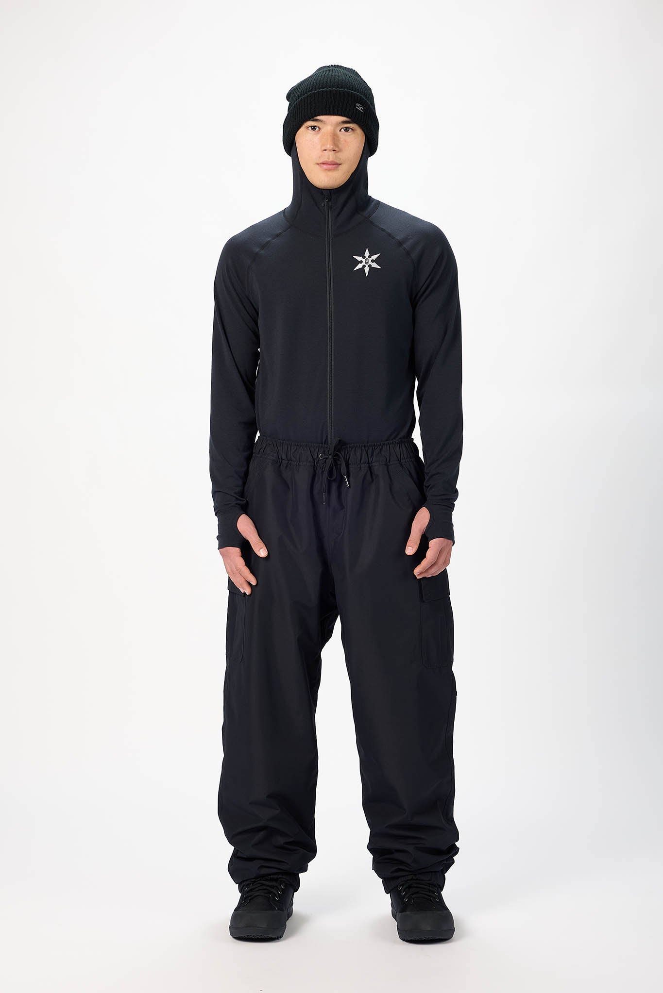 A model wearing a black hooded zip-up jacket and matching black pants with a small white logo on the chest, standing against a light gray studio background.