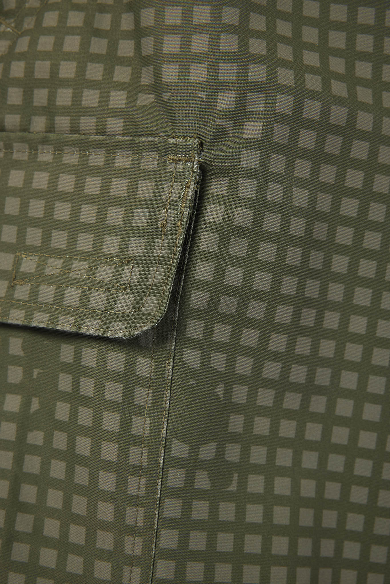 Close-up of a fabric surface with a small square grid pattern in muted sage green and off-white, showing part of a pocket flap and stitched seam.