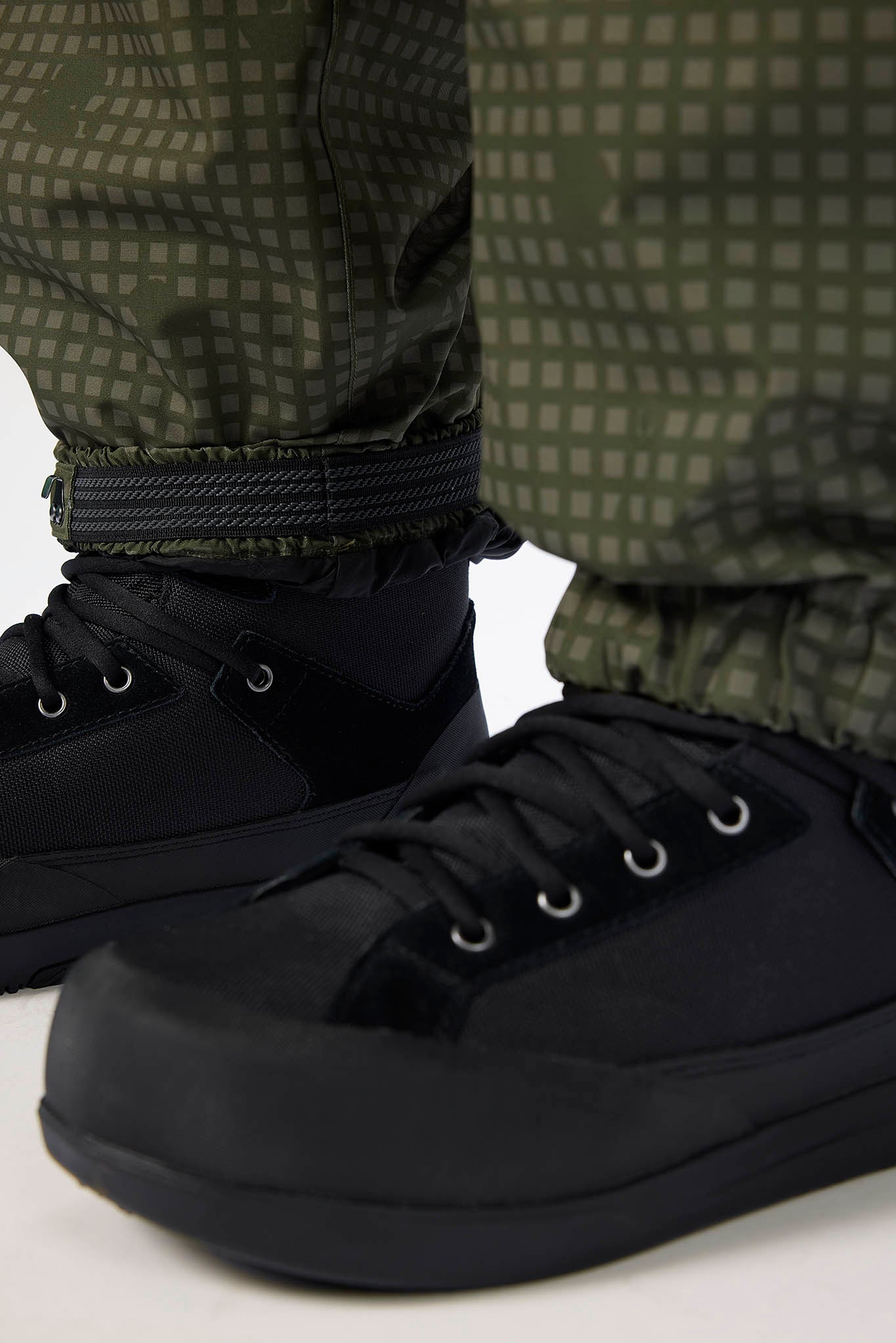 Close-up of a person wearing black low-top sneakers with black laces and olive green patterned pants, cropped to the lower legs and feet against a light studio background.