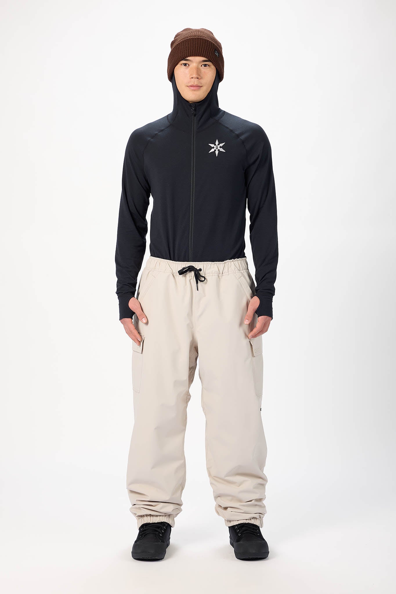 A male model wearing a black full-zip hooded jacket with a white logo on the chest, beige drawstring pants, black shoes, and a brown beanie, standing against a light gray studio background.
