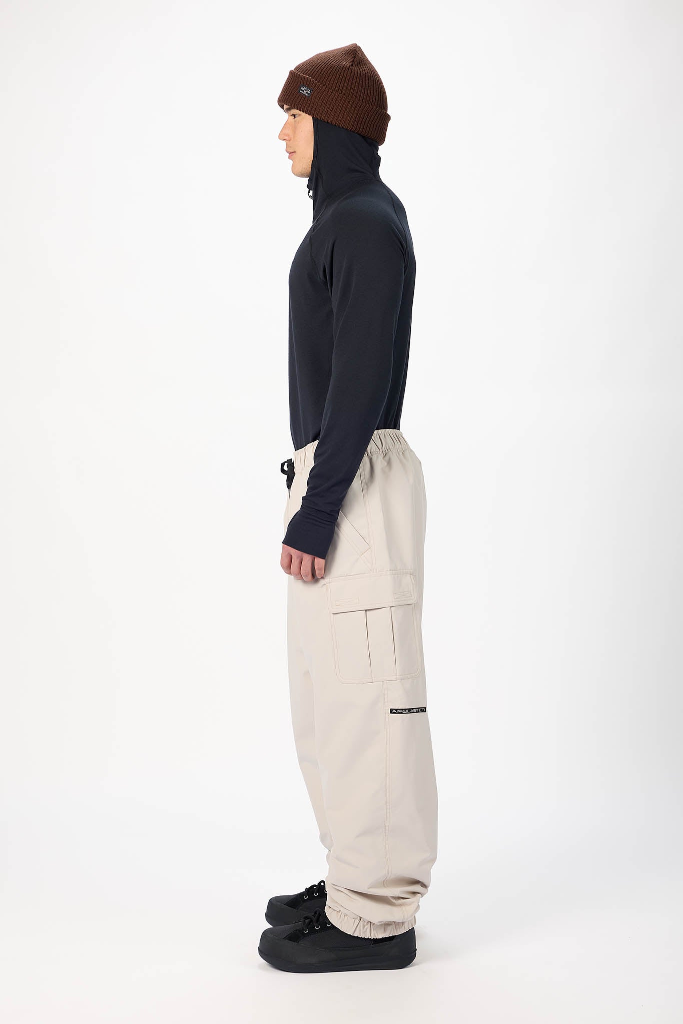 A model shown in profile wearing a dark navy long-sleeve turtleneck, beige cargo pants, black shoes, and a brown beanie against a plain white studio background.