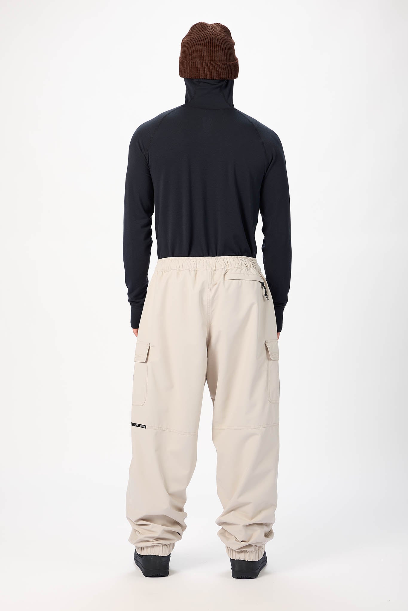 A person shown from the back wearing a dark navy long-sleeve turtleneck, beige cargo-style pants, and a brown knit beanie against a plain light gray studio background.