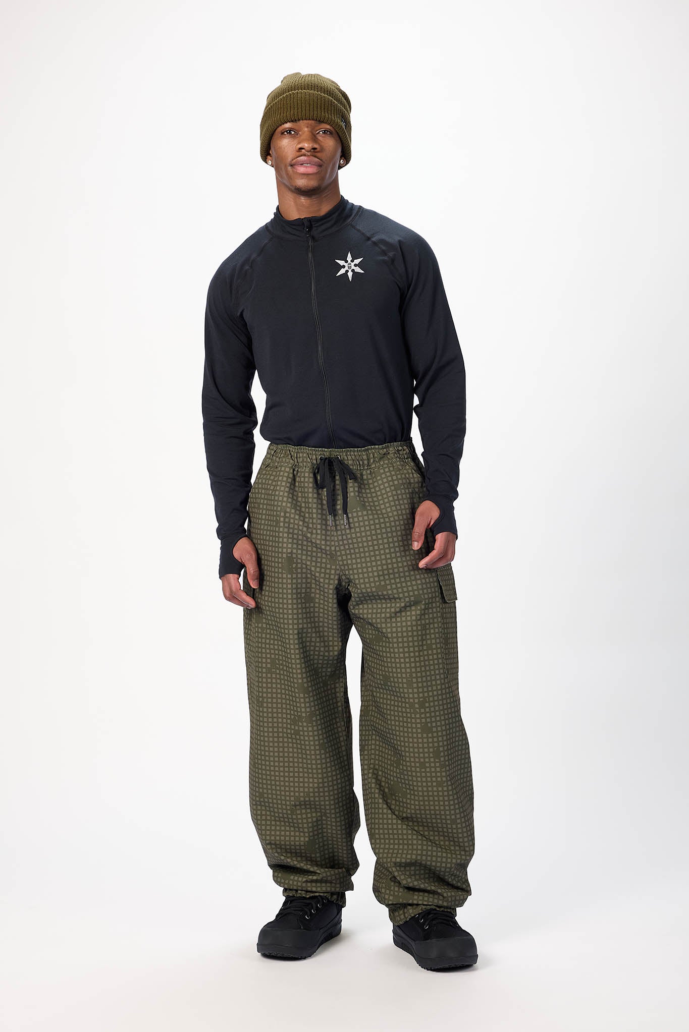 A man modeling a black long-sleeve athletic top with a white starburst logo on the chest, paired with olive green patterned pants, a matching olive beanie, and black sneakers, against a plain white studio background.