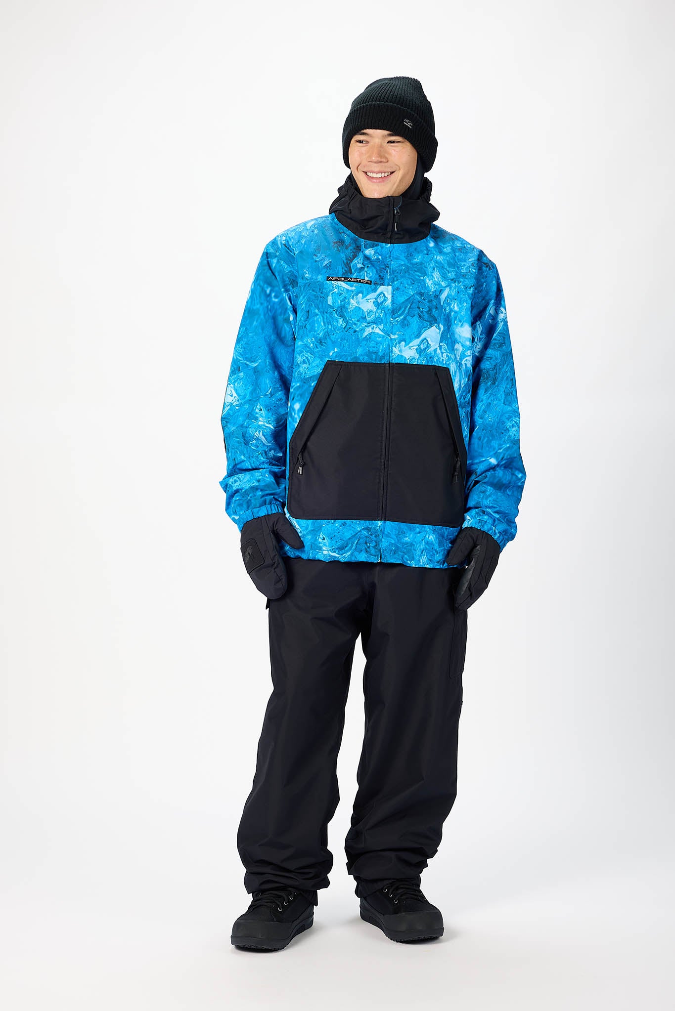 A model wearing a blue and black patterned snow jacket, black snow pants, a black beanie, and black gloves, standing against a light gray studio background.