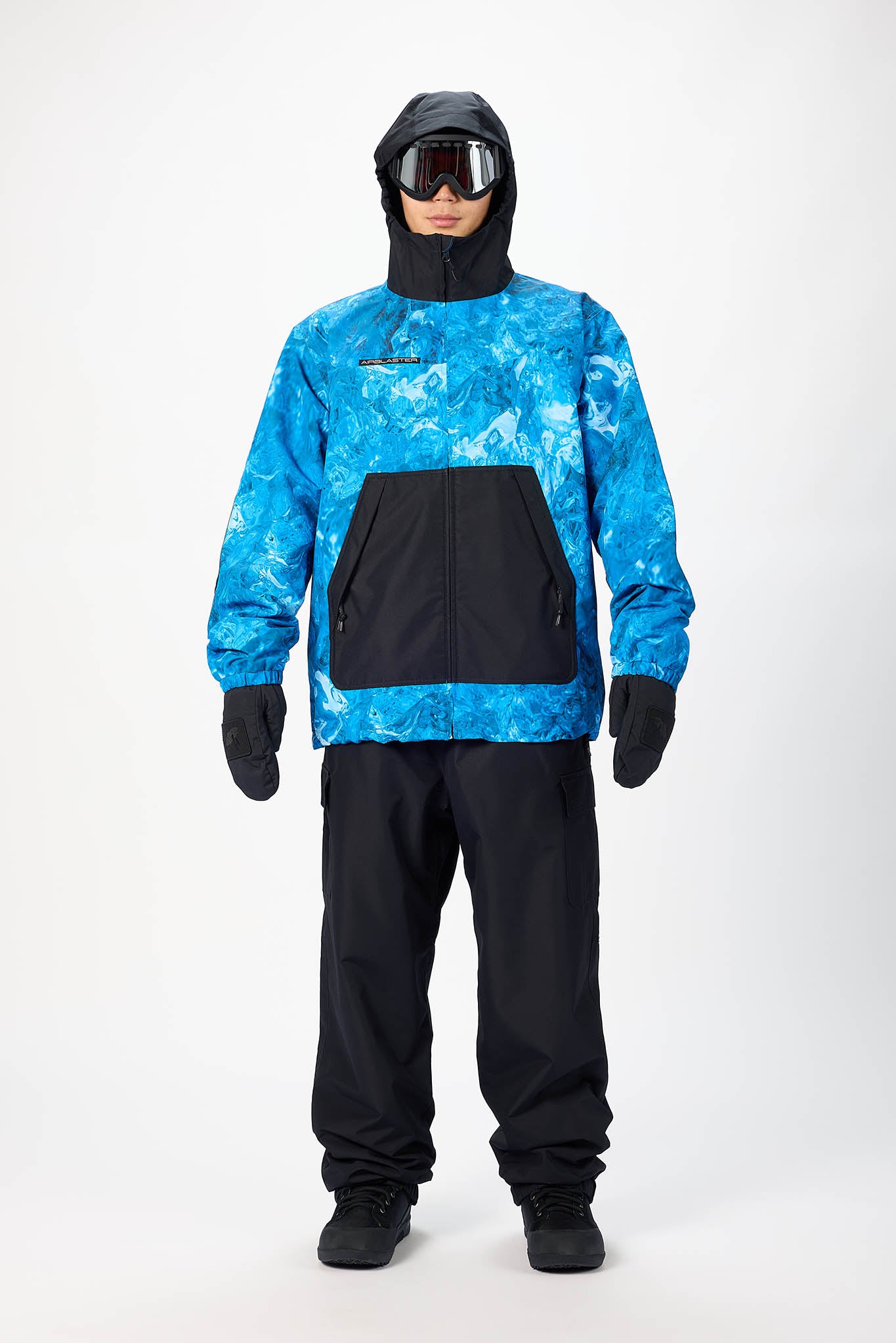 A model wearing a blue and black patterned ski jacket with a black hood, black snow pants, black gloves, and ski goggles, standing against a light gray studio background.