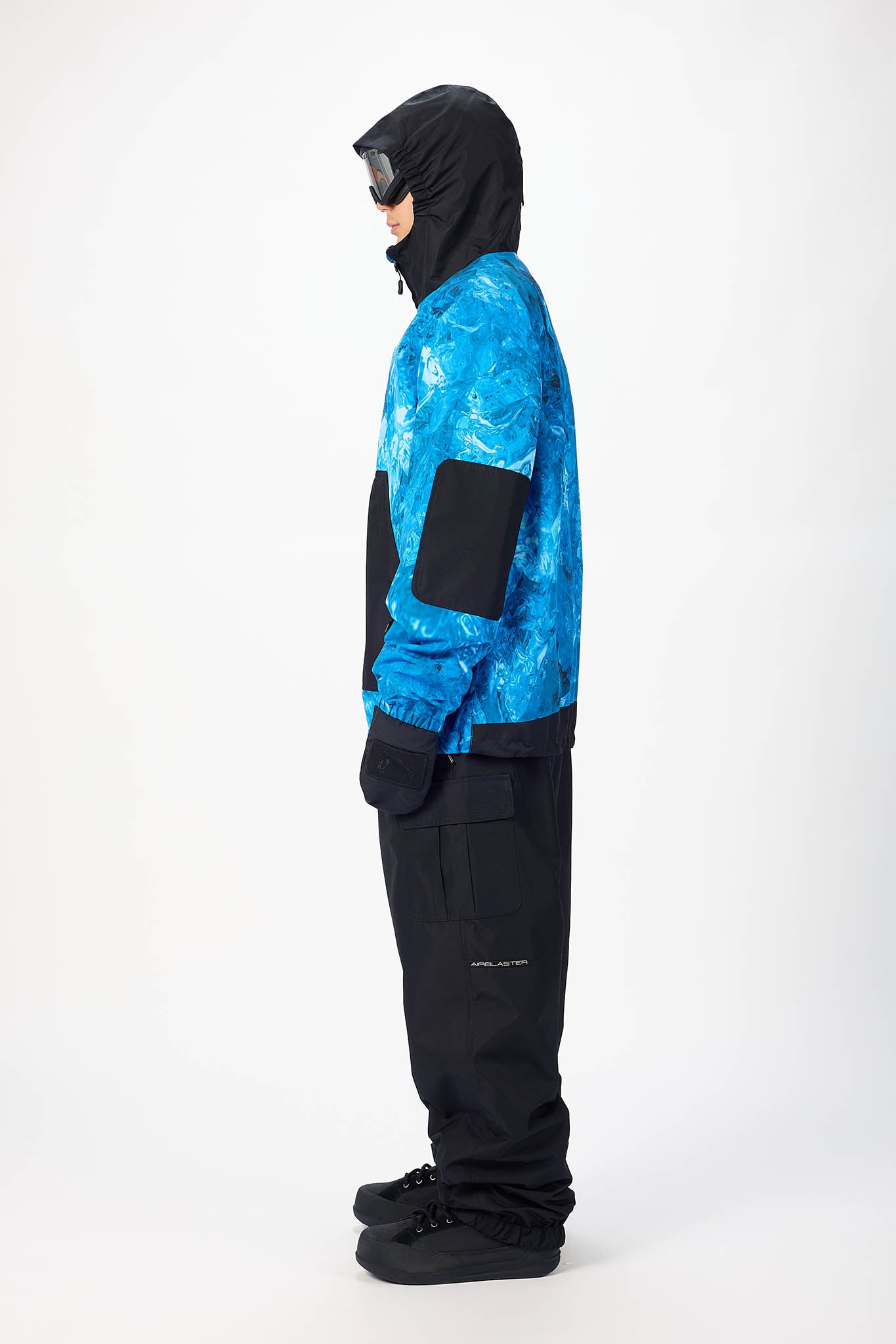 A person shown in profile wearing a blue and black patterned jacket with black pants, black hooded mask, and black shoes, photographed against a plain light gray studio background.
