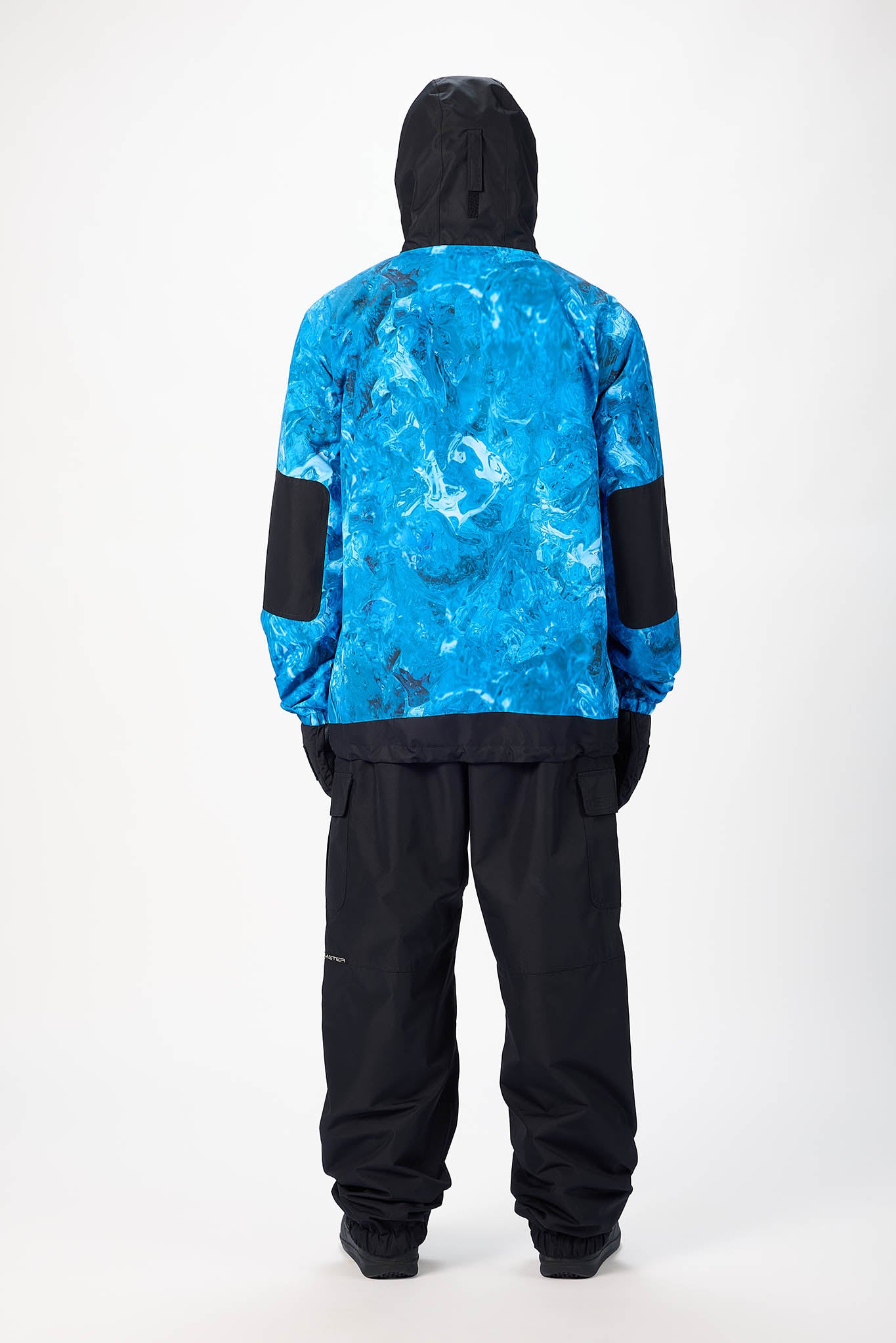 A person shown from the back wearing a hooded jacket with a bright blue marbled pattern and black sleeves, paired with black pants, against a plain light gray studio background.