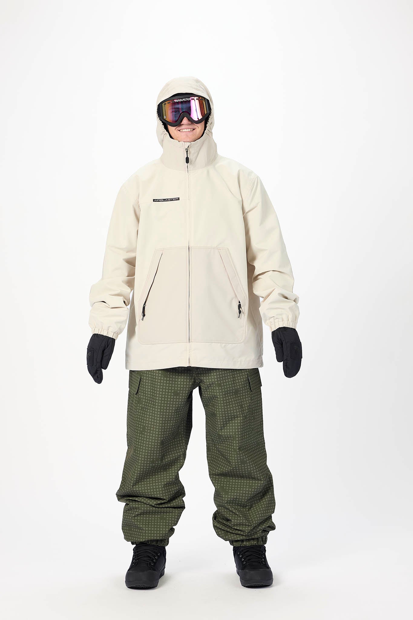 A model wearing a light beige hooded snowboard jacket, olive green patterned snow pants, black gloves, and black boots, standing against a plain white studio background.