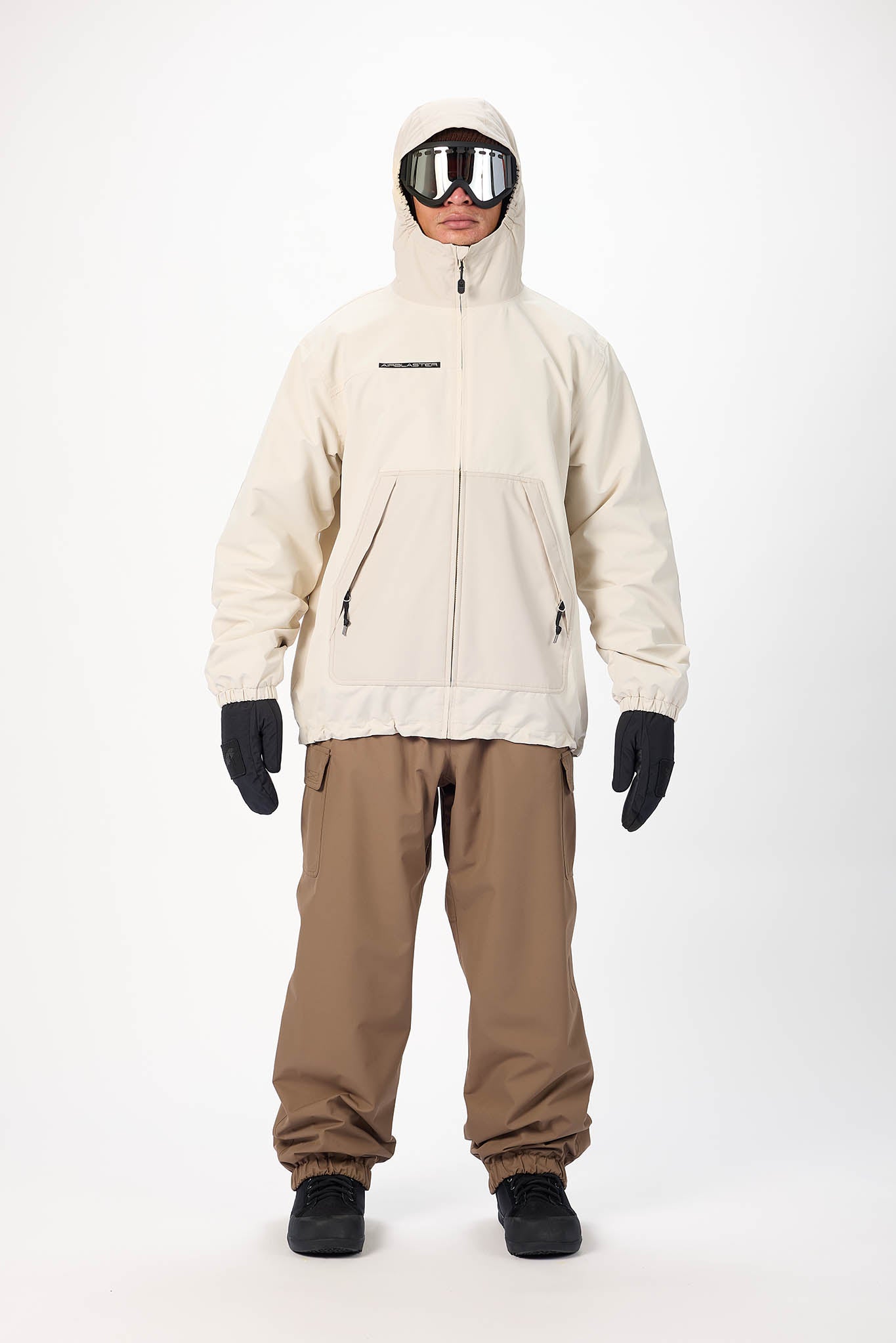 A model wearing a light beige hooded snowboard jacket, brown snow pants, black gloves, and black boots, standing against a plain white studio background.