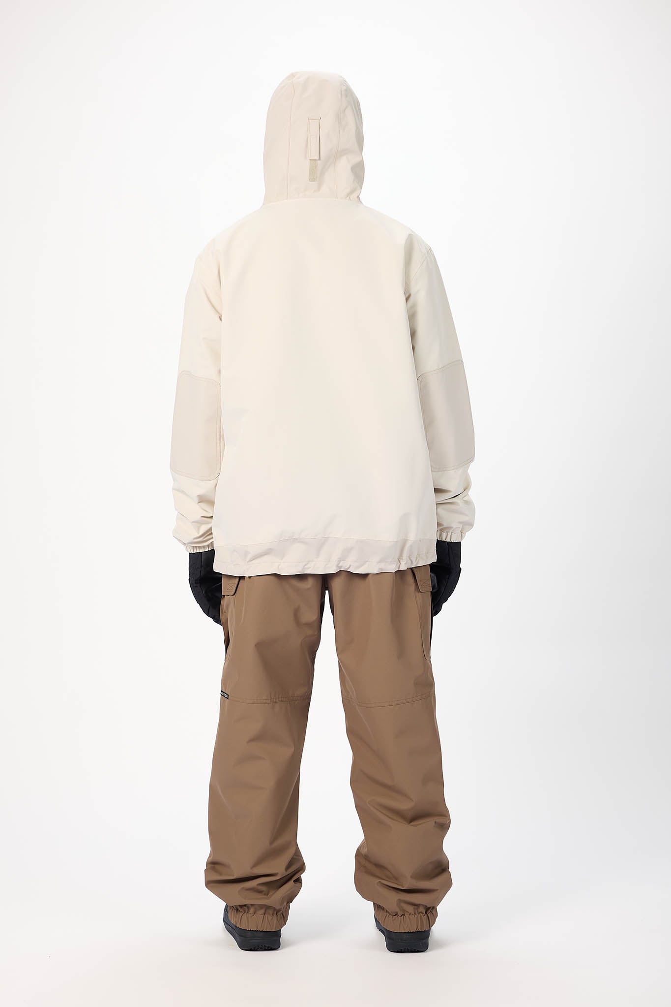 A person shown from the back wearing a light cream hooded jacket, brown snow pants, and black gloves against a plain white studio background.