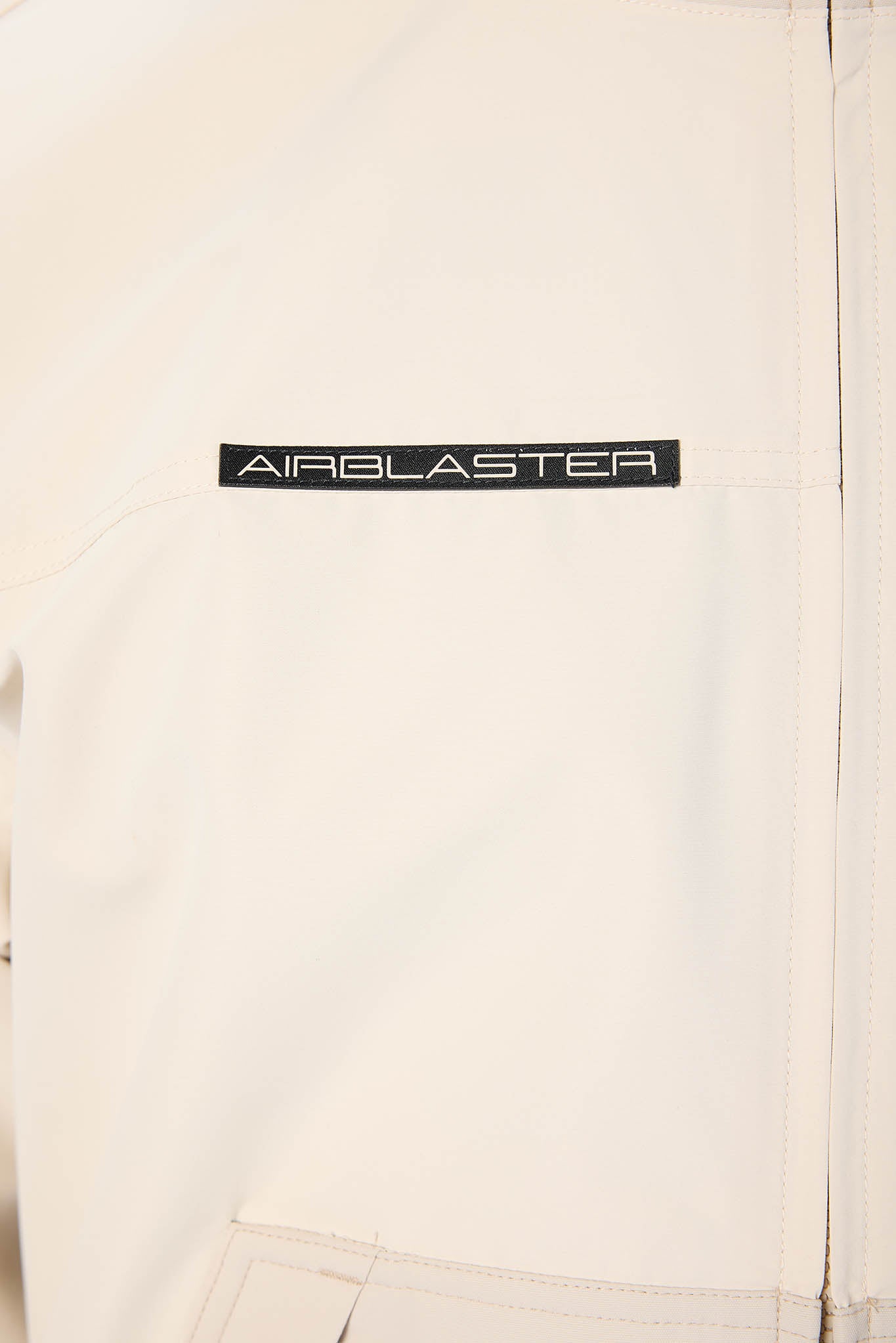 Close-up of a light cream-colored jacket featuring a black rectangular patch with the brand name 'AIRBLASTER' in white text.