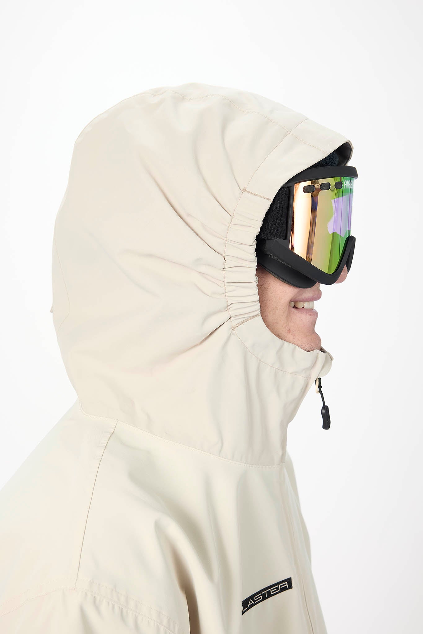 A close-up side profile of a person wearing a light beige hooded snowboard jacket and black ski goggles with mirrored lenses, against a plain white background.