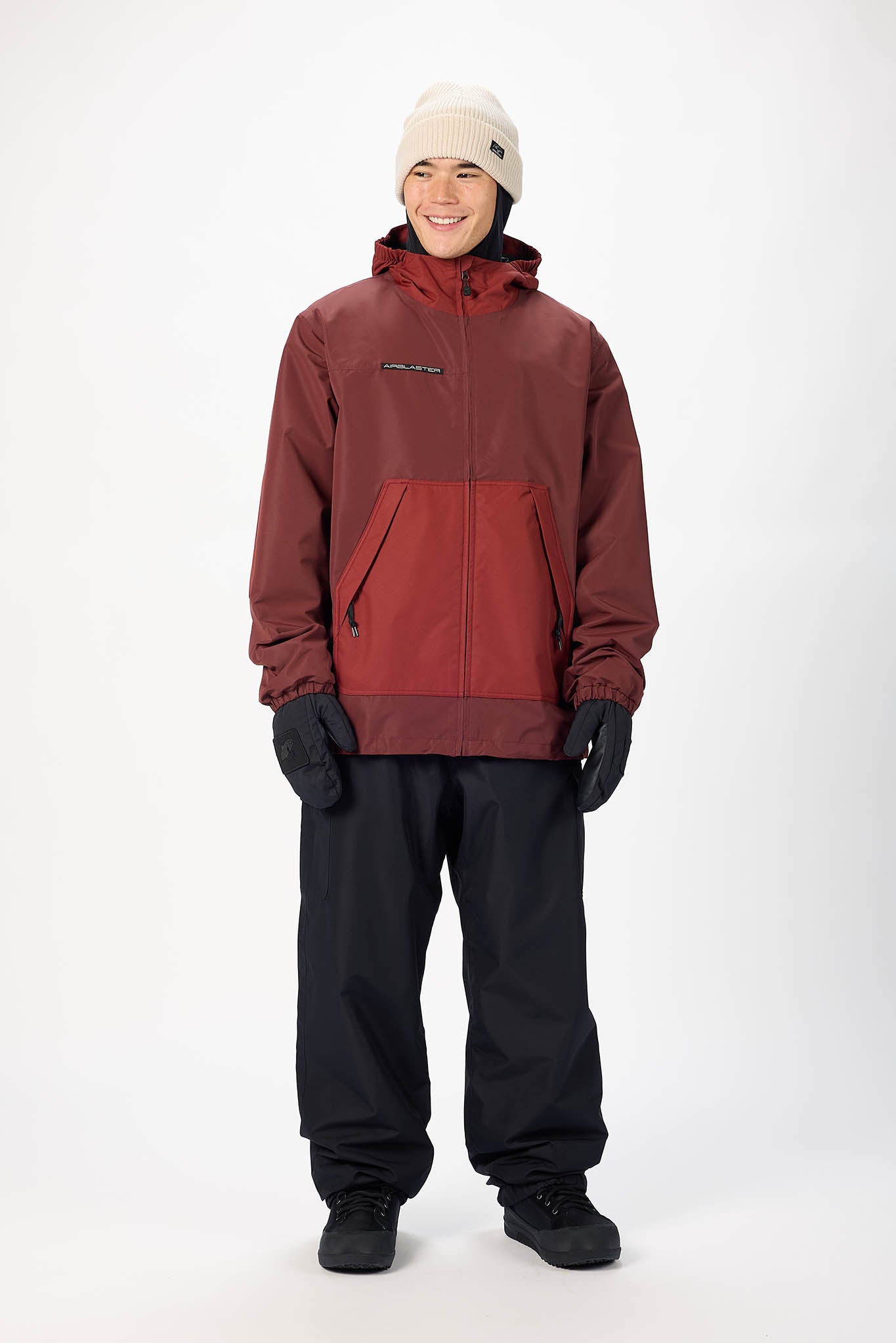 A man modeling a two-tone red and maroon hooded snowboard jacket, paired with black snow pants, black gloves, and a cream beanie, against a plain white studio background.