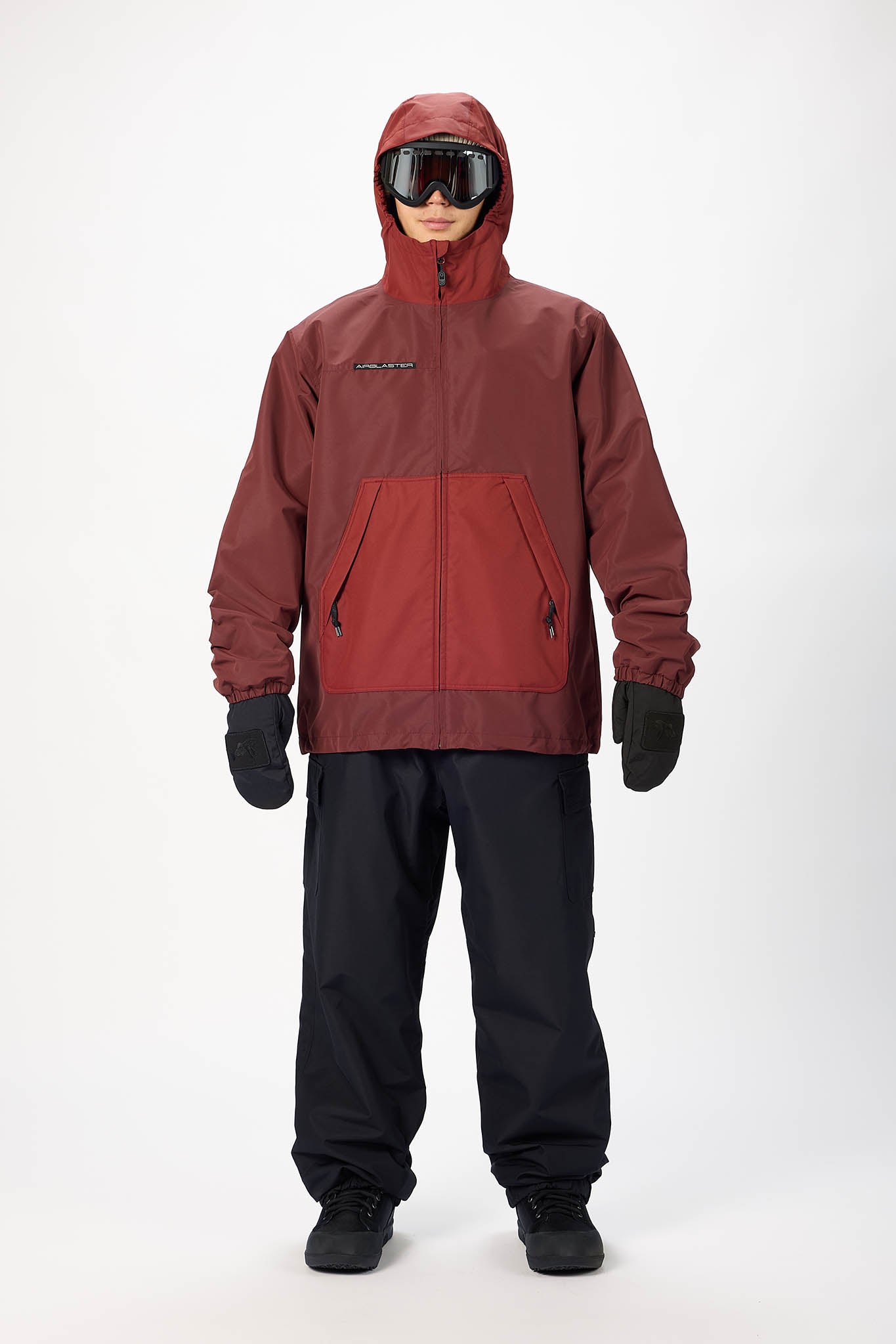 A model wearing a hooded ski jacket in a dark red and rust color-blocked design, paired with black snow pants, gloves, and goggles, standing against a plain white studio background.